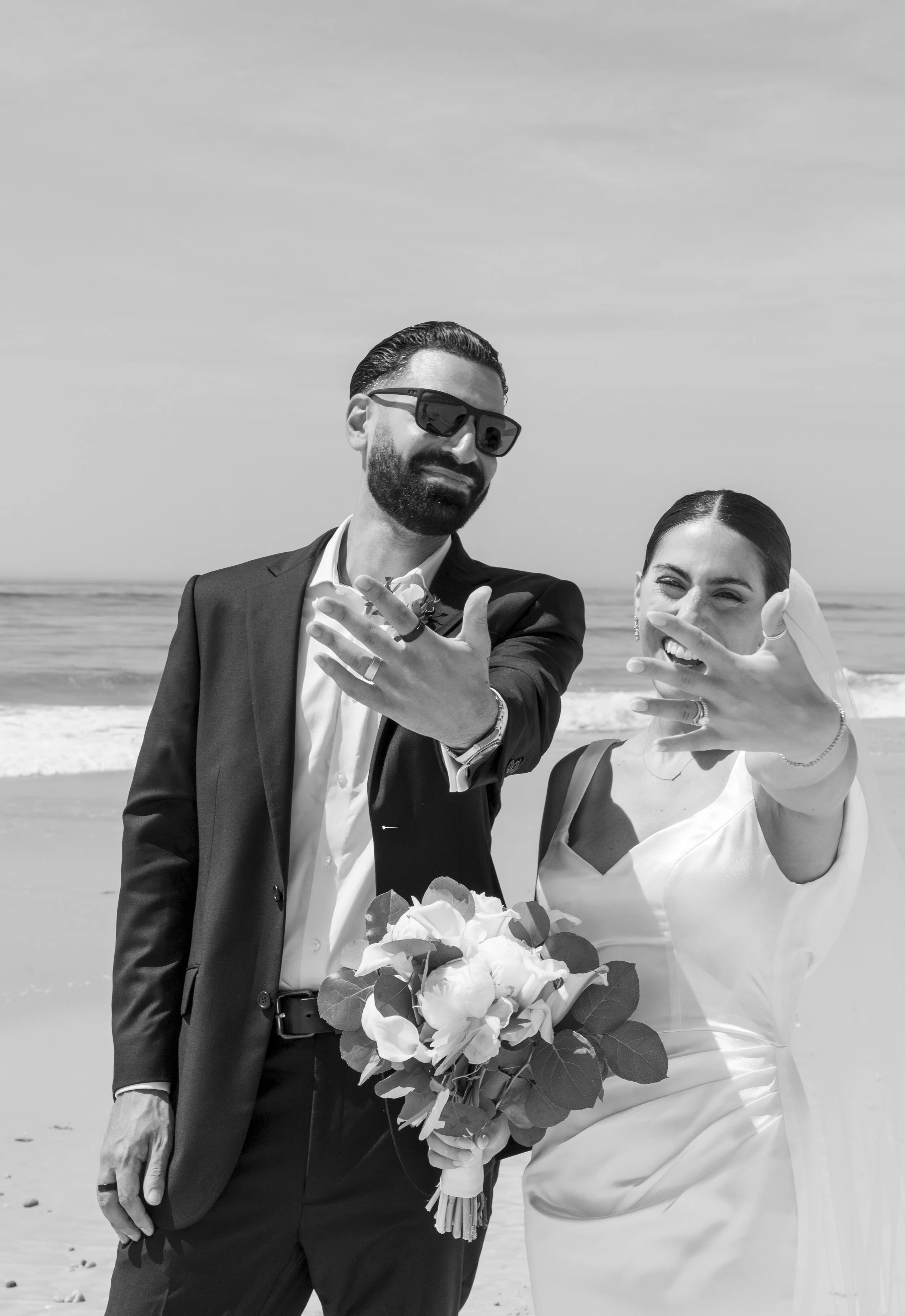A wedding couple on the beach, with the bride holding a bouquet of flowers, both showing off their wedding rings, and the groom wearing sunglasses.