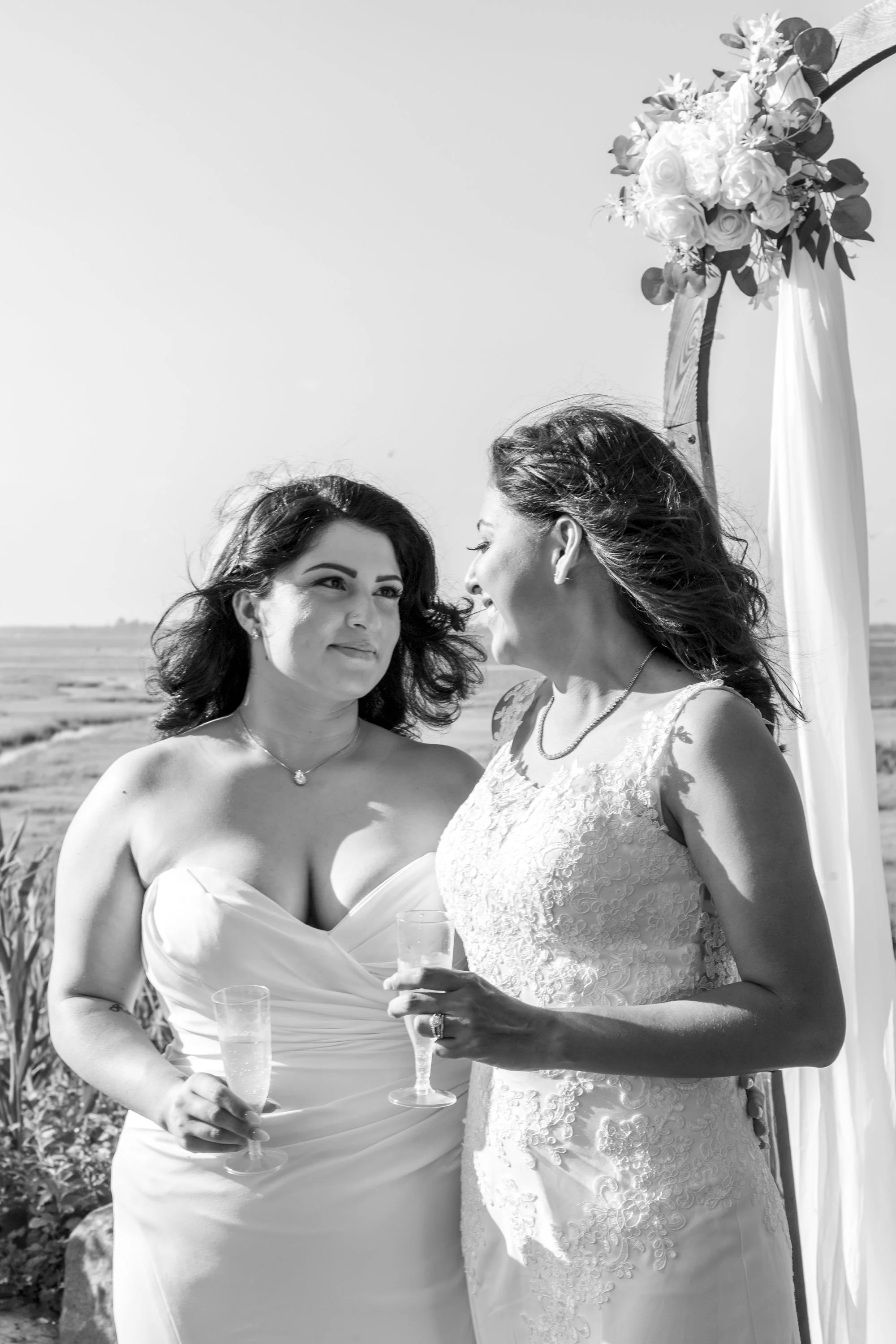 Two women in wedding dresses standing outdoors, holding champagne glasses, with an archway decorated with flowers behind them.