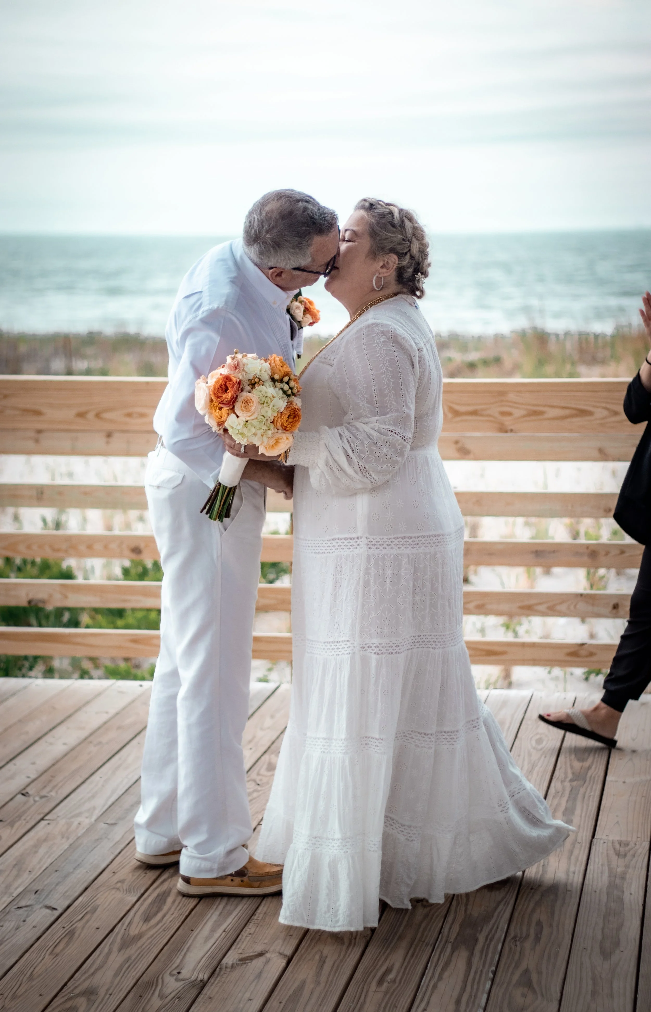A couple is kissing during their wedding on a wooden deck near the beach, with a bouquet of peach, orange, and white flowers, and ocean in the background.