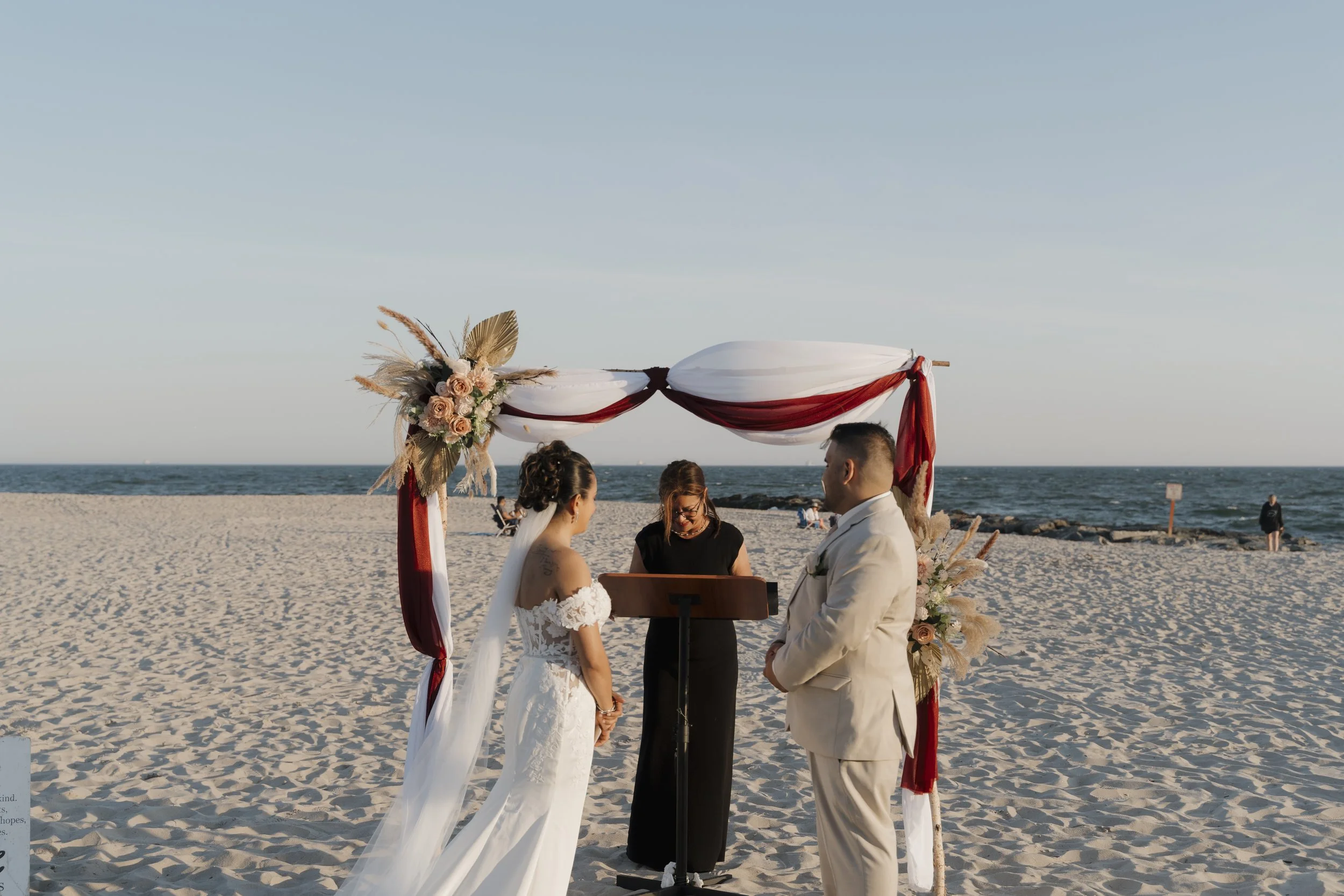 A wedding ceremony taking place on a beach with an arch decorated with flowers, pampas grass, and draped fabric, as the bride and groom stand facing each other under the arch with an officiant.