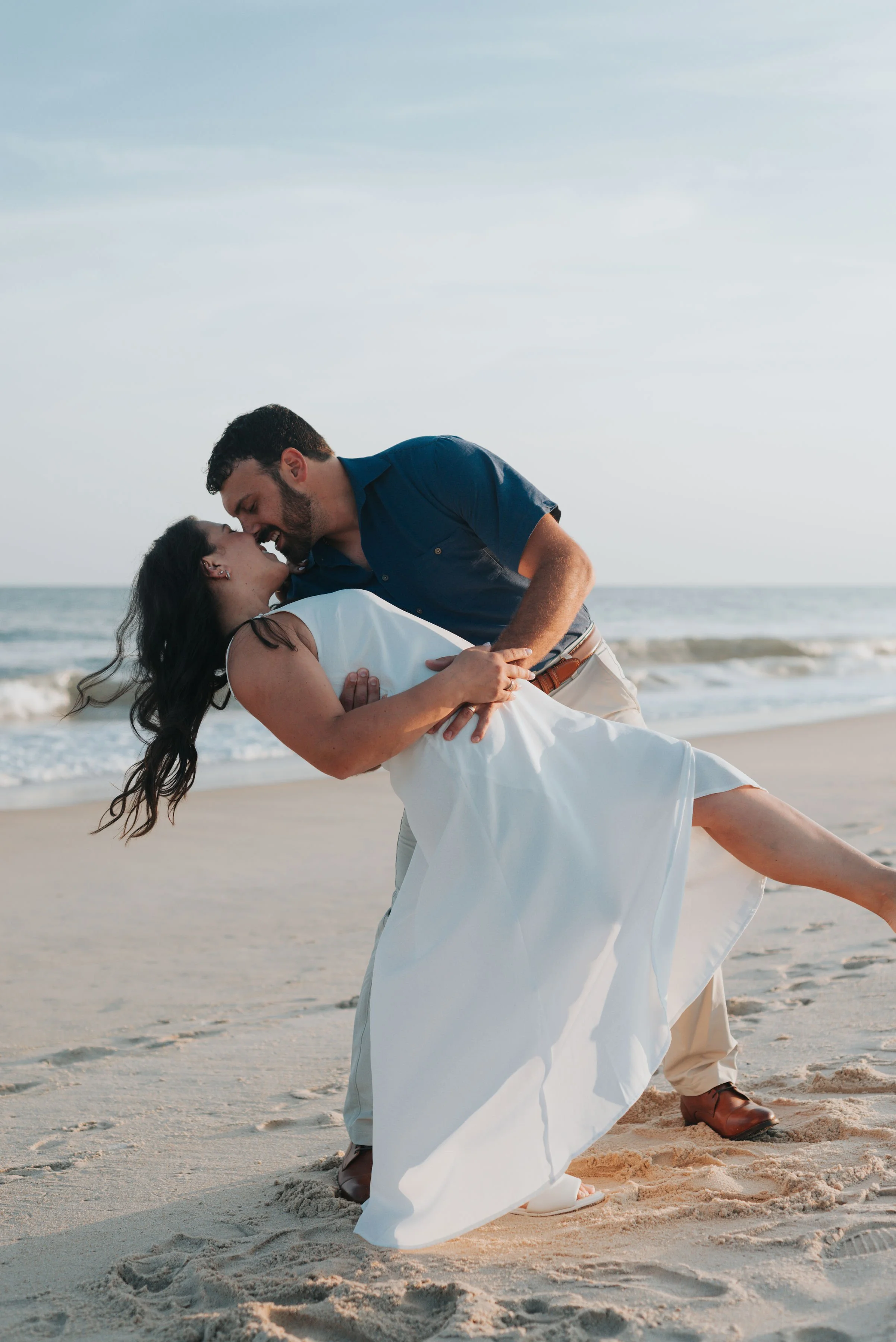 A couple sharing a romantic moment on the beach, with the man dipping the woman, both smiling, during sunset or late afternoon.