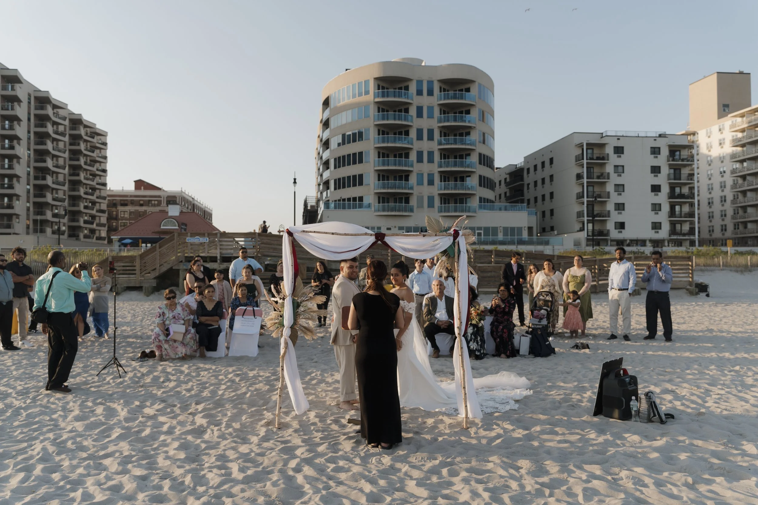 A beach wedding ceremony with a couple standing under a decorated wooden arch, surrounded by wedding guests sitting and standing on the sandy beach, with modern buildings in the background.