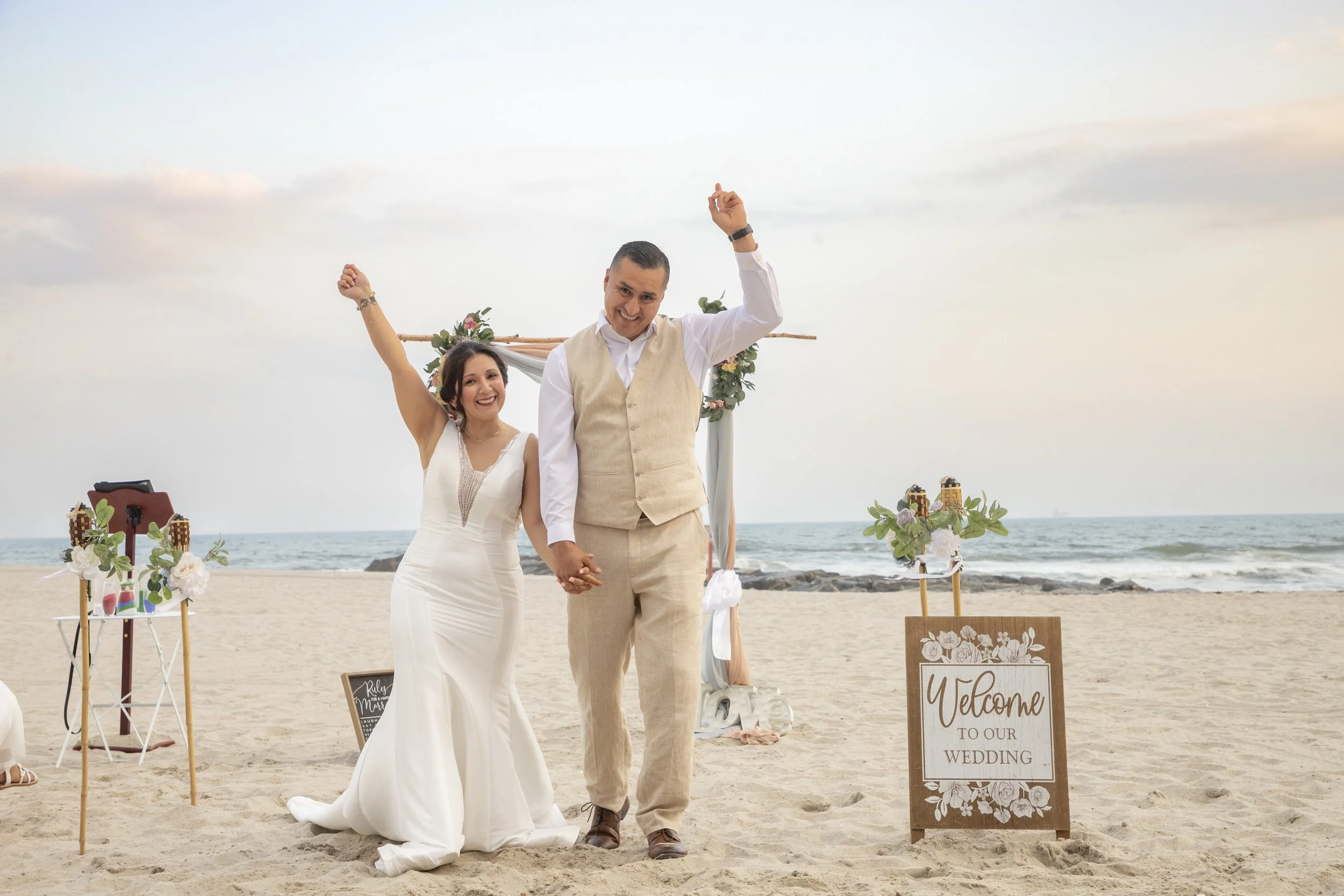Happy couple in wedding attire holding hands and celebrating on the beach with a wooden welcome sign and floral decoration in the background.