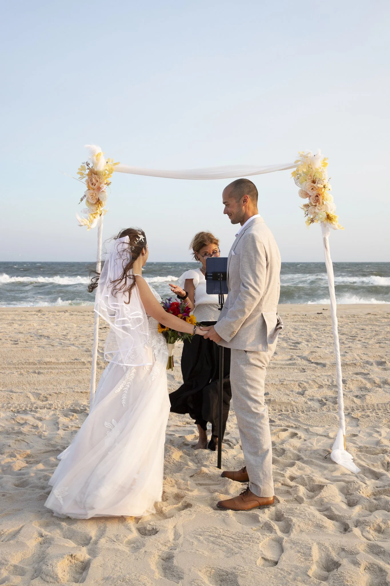 A beach wedding ceremony with a couple exchanging vows under a floral arch, officiant present, ocean in the background, sand beneath their feet.