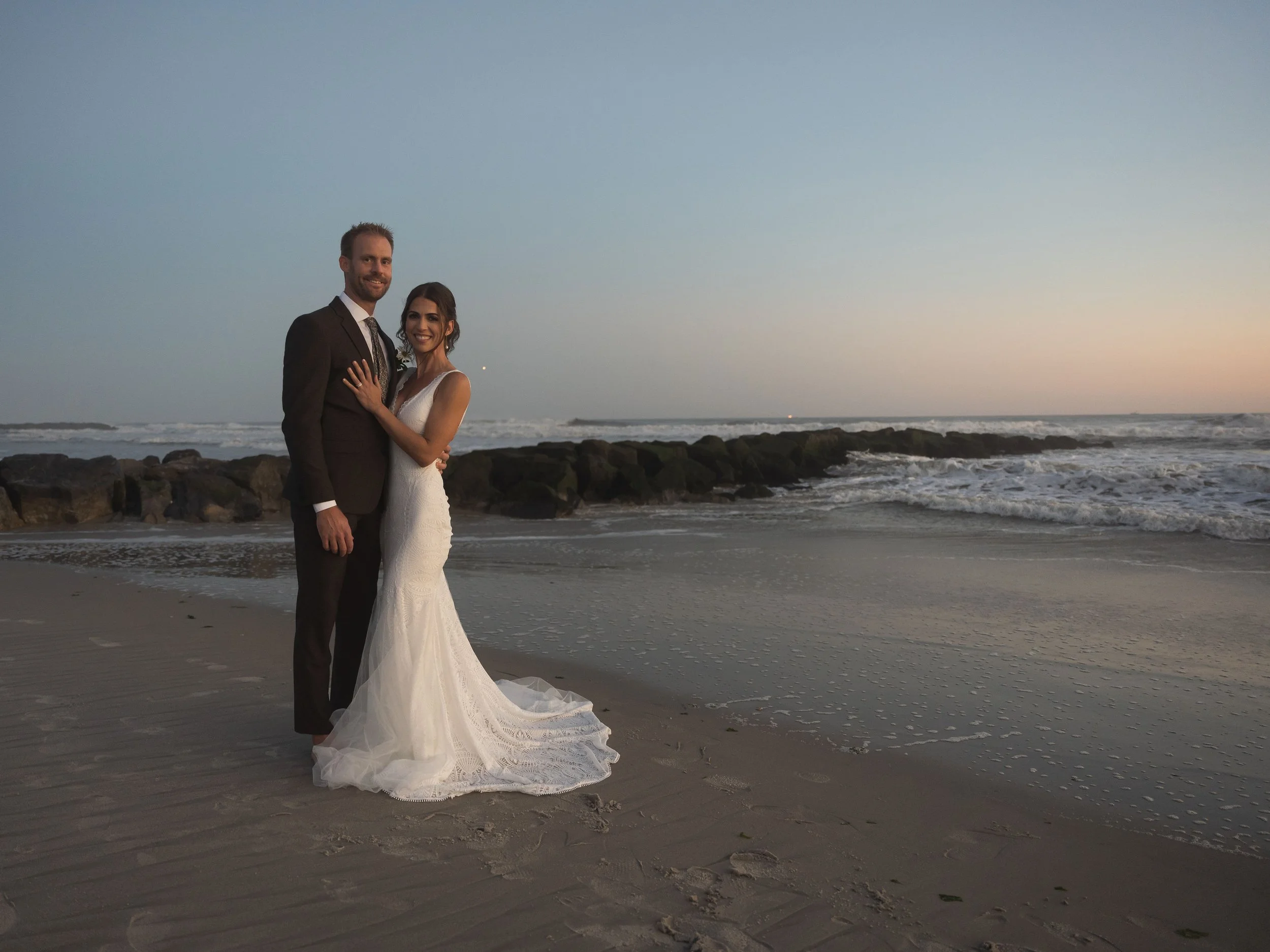 A newlywed couple in wedding attire stands on the beach at sunset, with rocks and ocean waves in the background.
