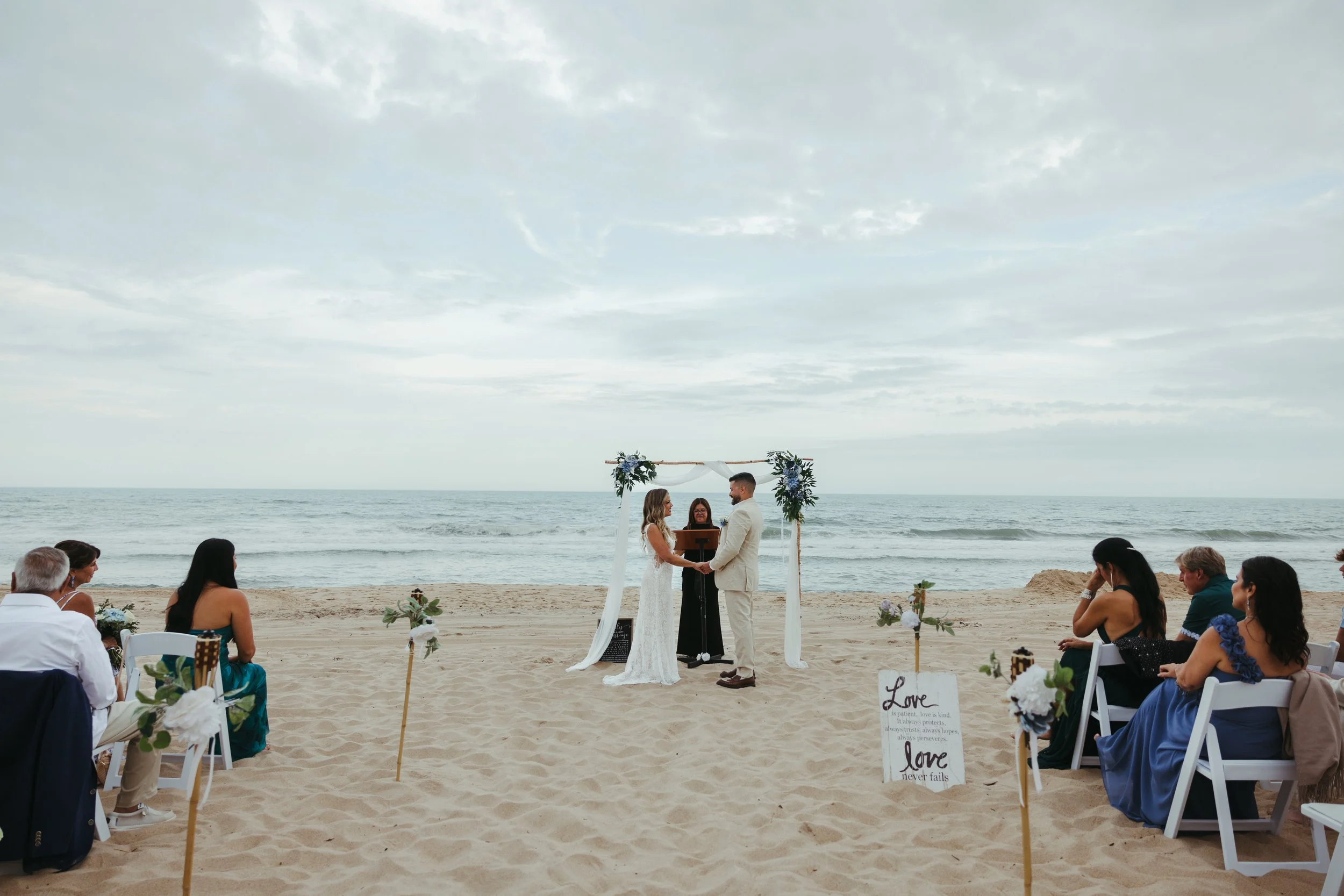 Beach wedding ceremony with a couple exchanging vows under a floral arch, surrounded by seated guests facing the ocean, with a cloudy sky overhead.