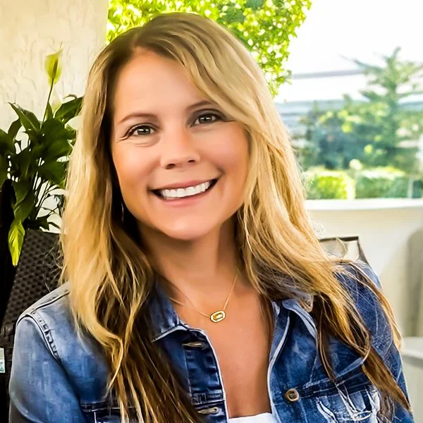 Smiling woman with blonde hair wearing a denim jacket, outdoors with greenery in the background.