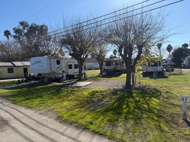 A small campground with several parked RVs or mobile homes on a grassy area, with trees, and a clear blue sky.