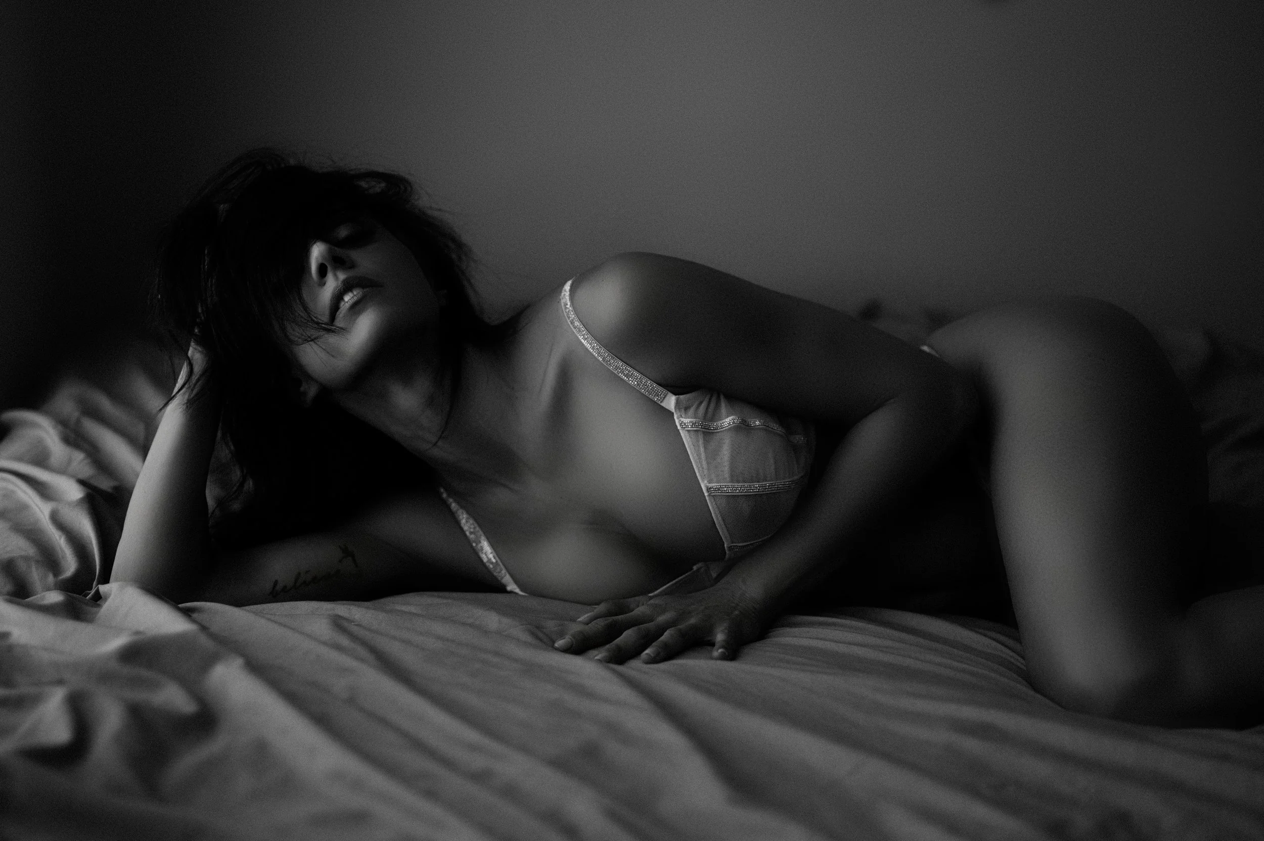 A black and white photo of a woman lying on a bed, wearing lingerie, with her hair partially covering her face, in a moody, intimate setting.