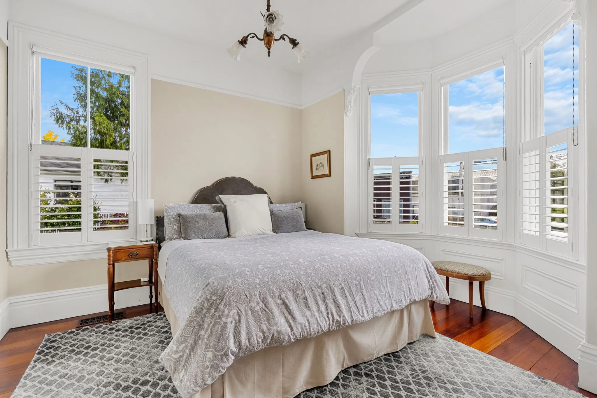 Bright bedroom with large window, floral wallpaper, bed with white linens and pillows, small sitting area with upholstered chair, small side table, and vintage wooden desk, hardwood floor, ceiling light fixture.