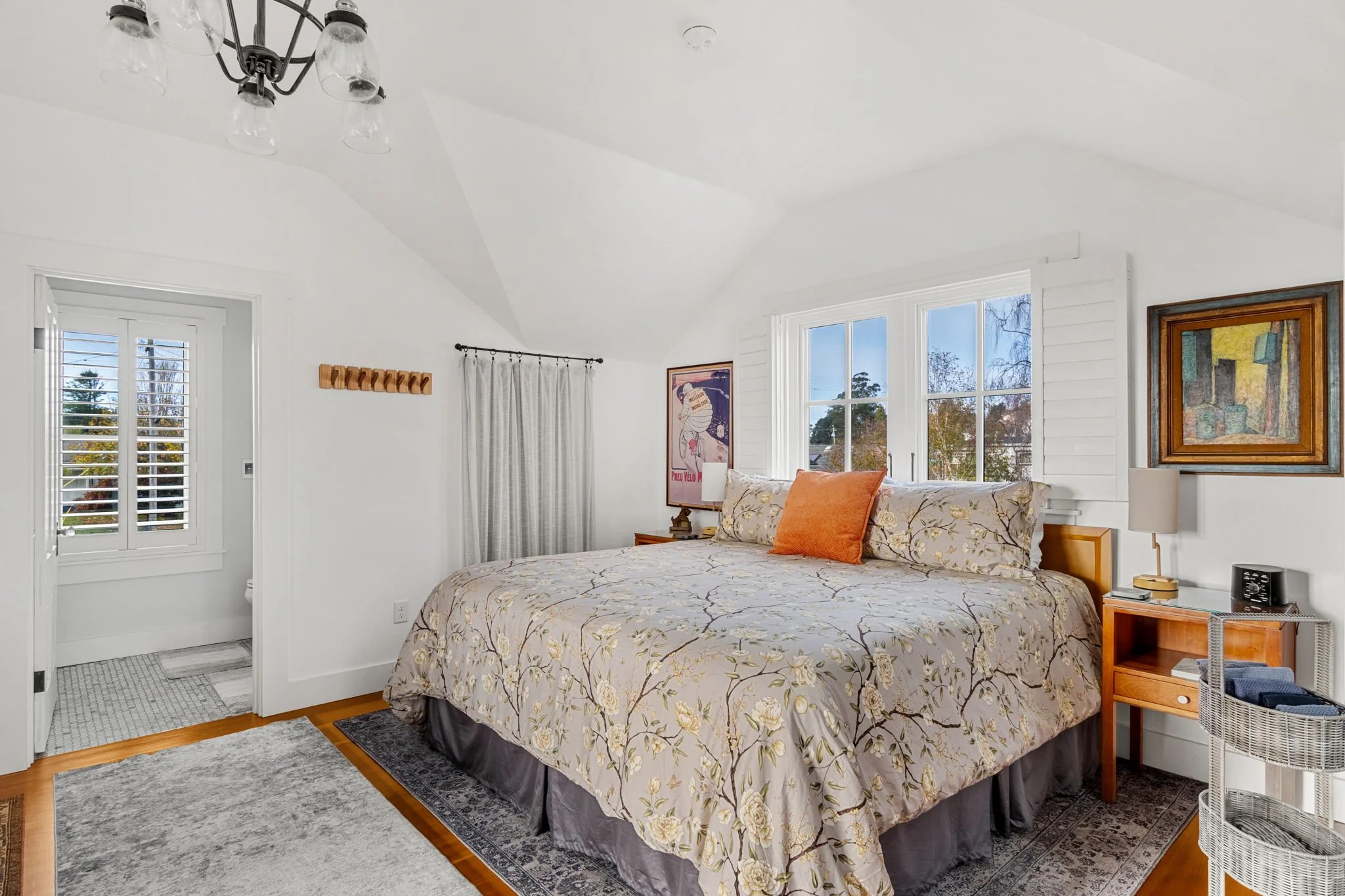 A bedroom with a large bed with white bedding and decorative pillows, a window with white shutters, a ceiling light fixture with multiple bulbs, a small nightstand with a black lamp, and a wicker side table near the bed.