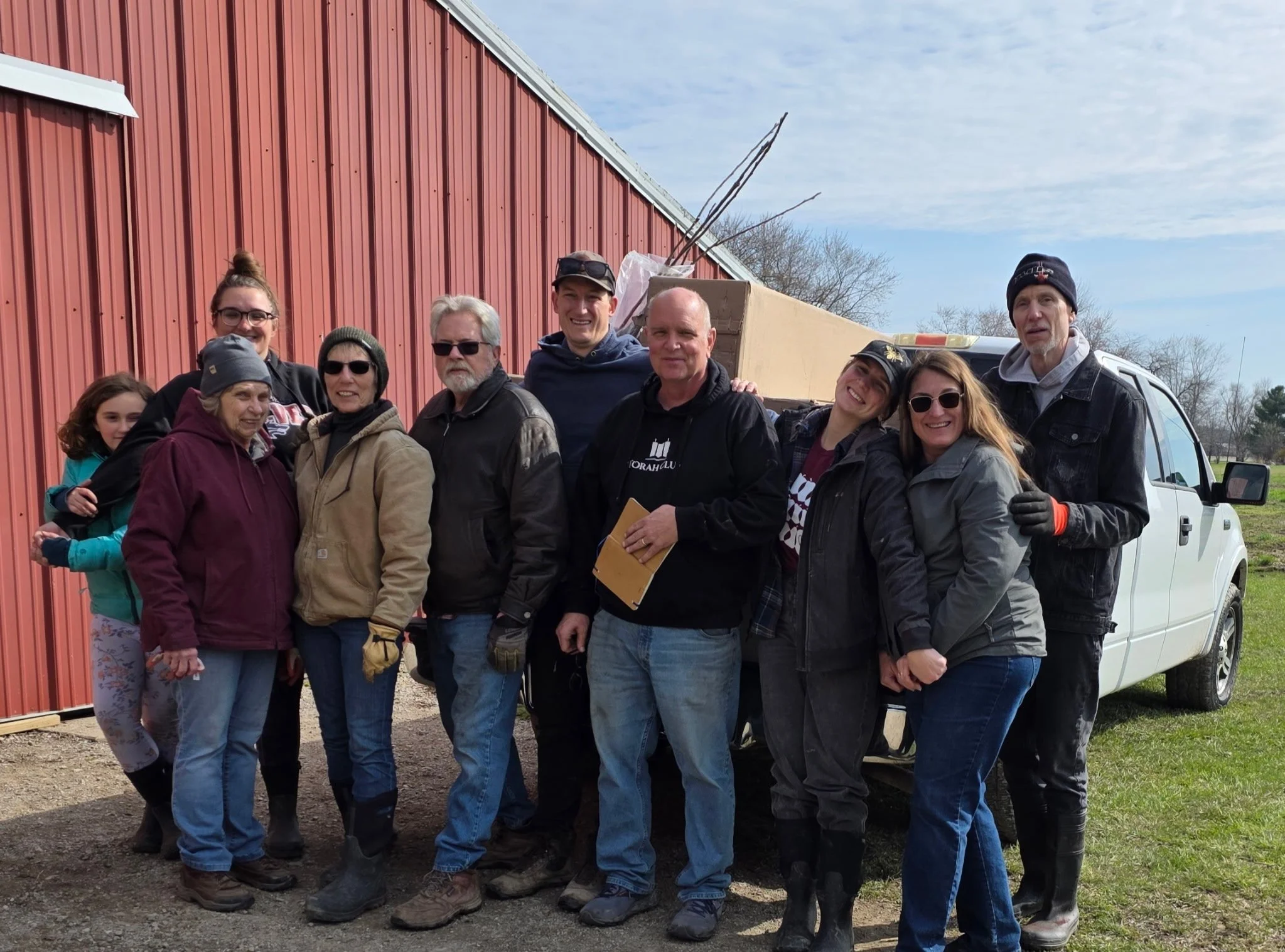 A group of people gathered outdoors in front of a red barn, smiling for the camera. The group includes men and women of various ages, wearing casual and work clothing, with some holding papers or tools. There is a white pickup truck parked nearby and a partly cloudy sky above.