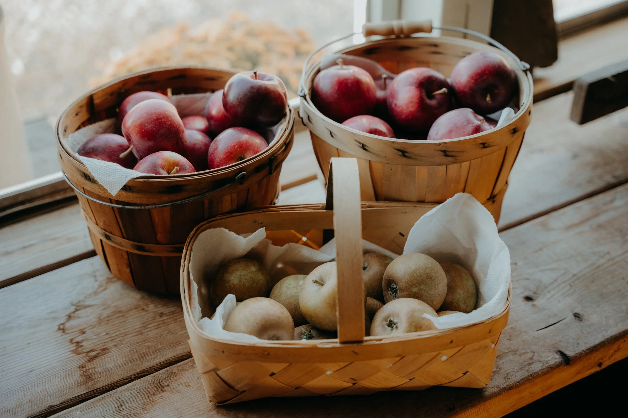 Three woven baskets filled with apples and pears on a rustic wooden table near a window.