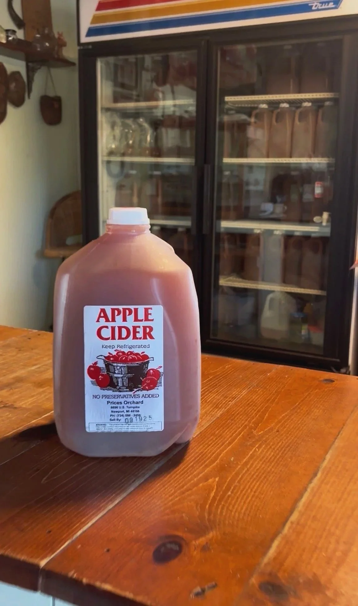 A bottle of apple cider on a wooden table in front of a refrigerator with glass doors inside a room.