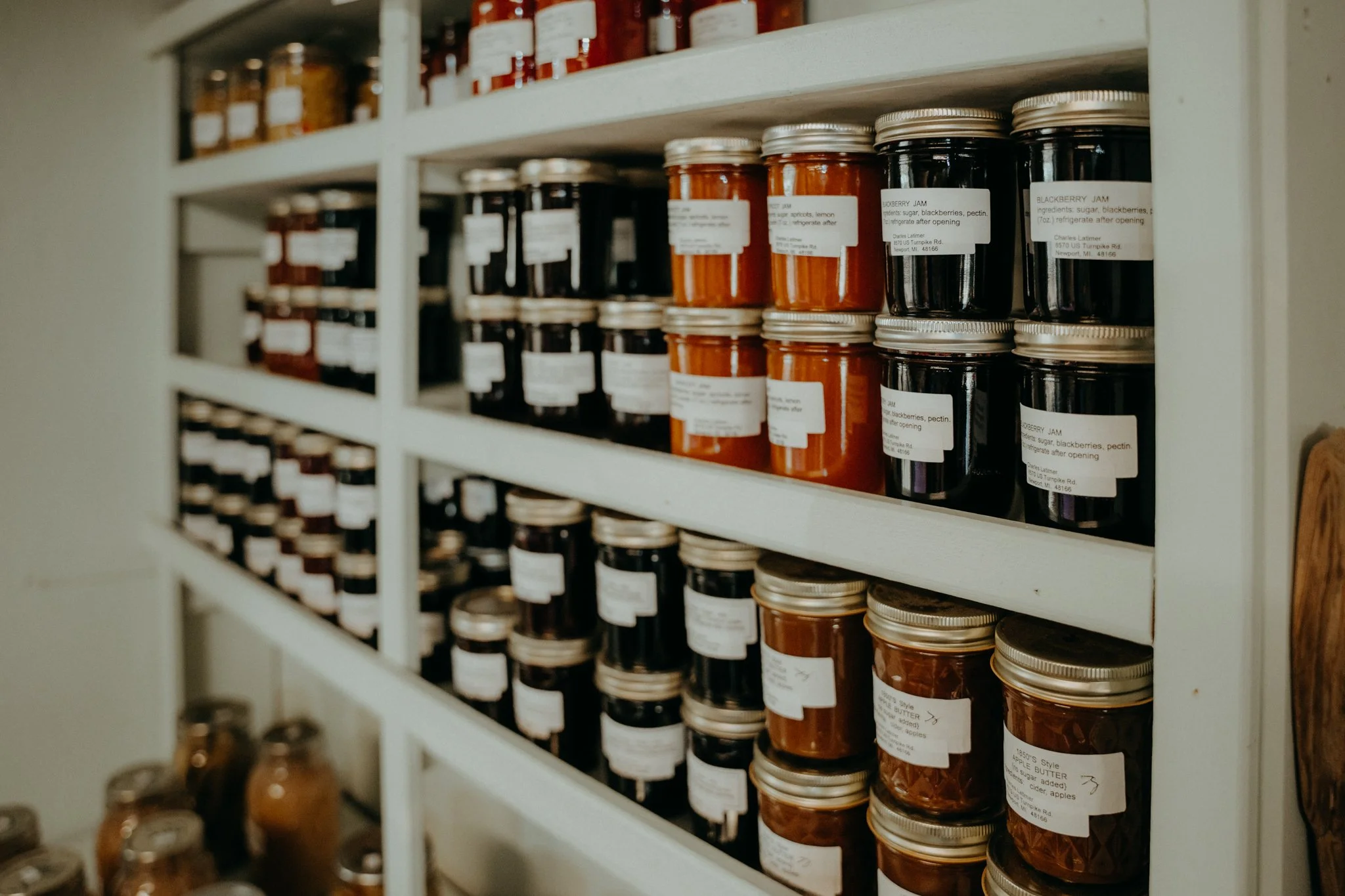 Close-up of a white wooden shelf filled with glass jars of homemade jams and preserves, each with white labels and silver lids.