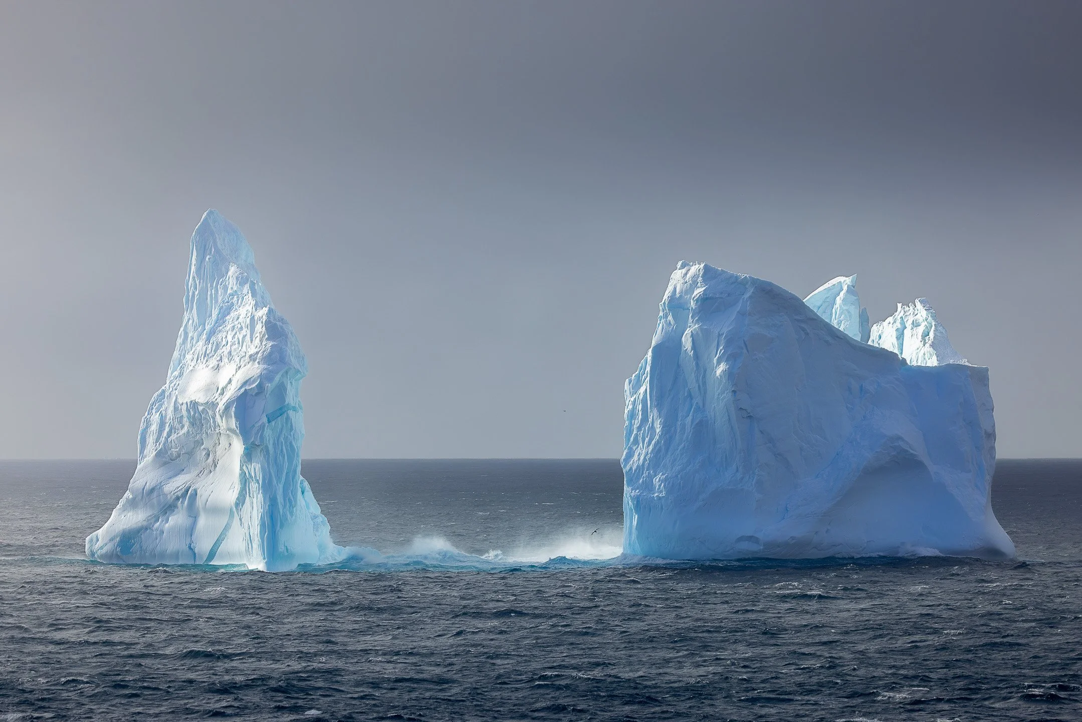 Two ice towers rise from a rough Southern Ocean sea near the Balleny Islands, catching directional light while waves break white at their base, with a seabird visible in the spray.