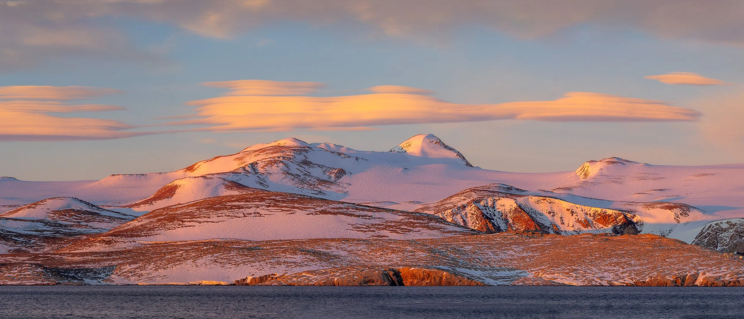 Golden morning light illuminates snow-dusted mountains at Terra Nova Bay and horizontal lenticular clouds above a dark blue sea.