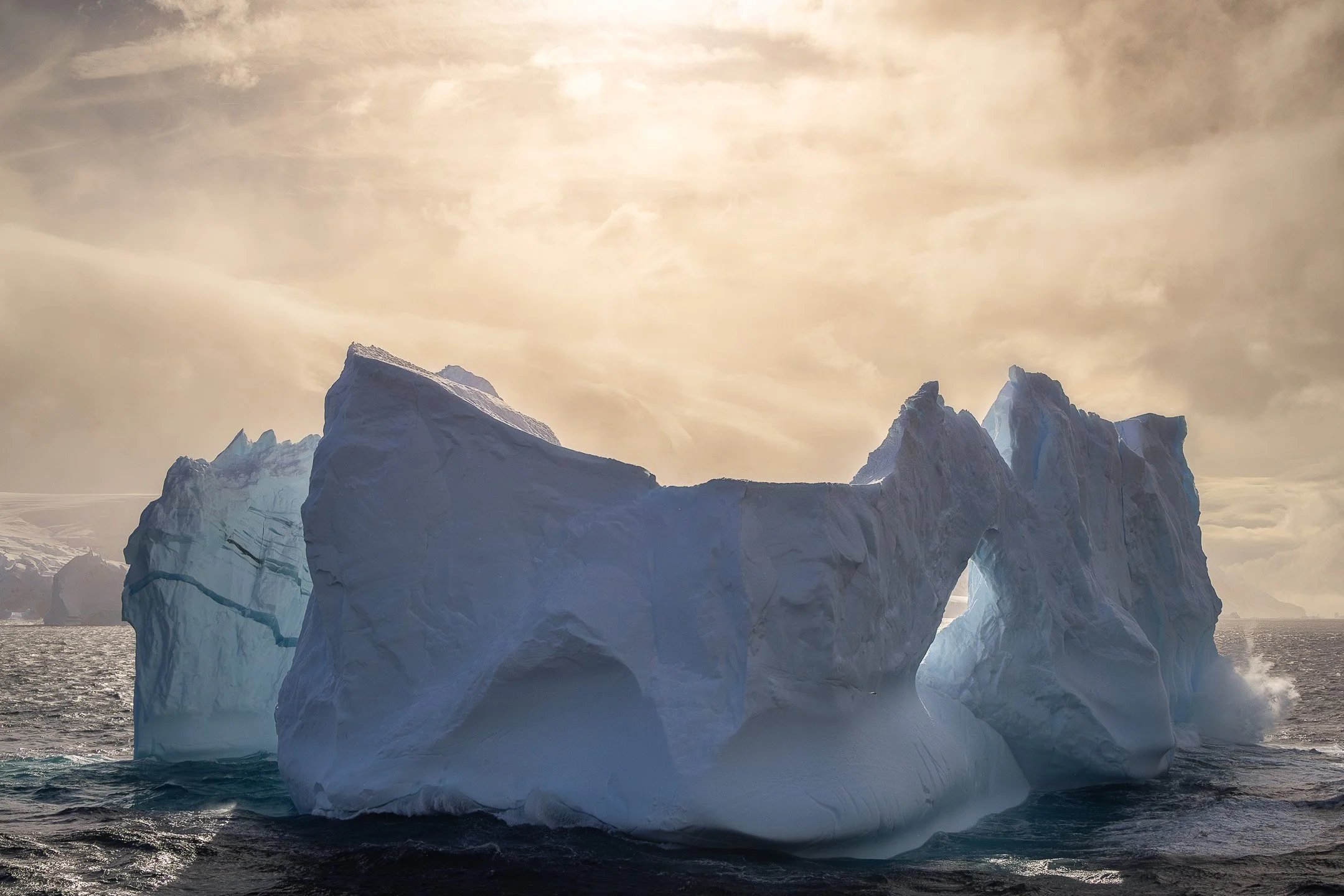 A monumental arch worn through the body of a large iceberg near the Balleny Islands, Antarctica, framing a golden sky beyond — one of the most extraordinary natural ice formations in the Southern Ocean.
