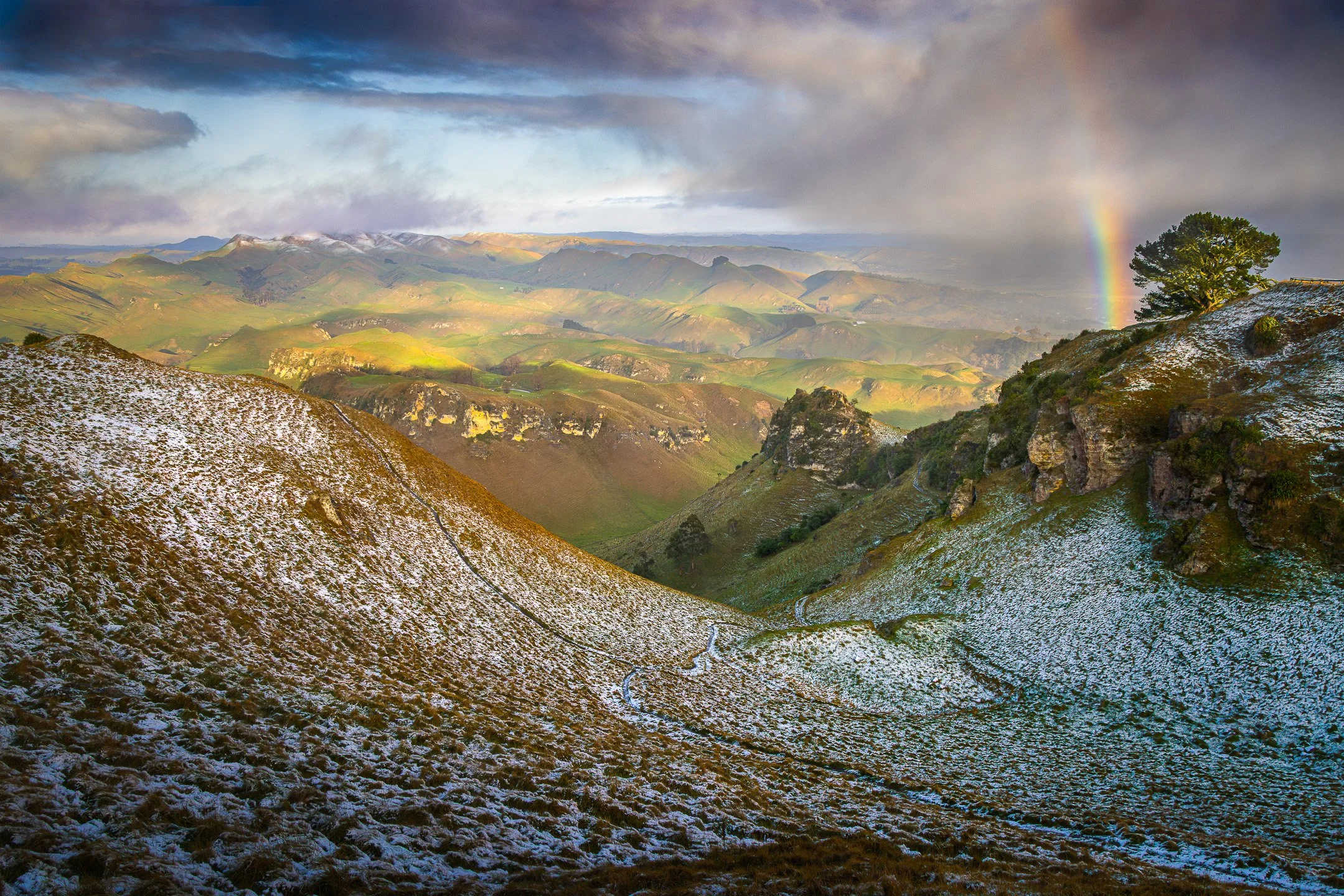 Snow-dusted valley on Te Mata Peak with a rainbow arching over the ridgeline at sunrise.