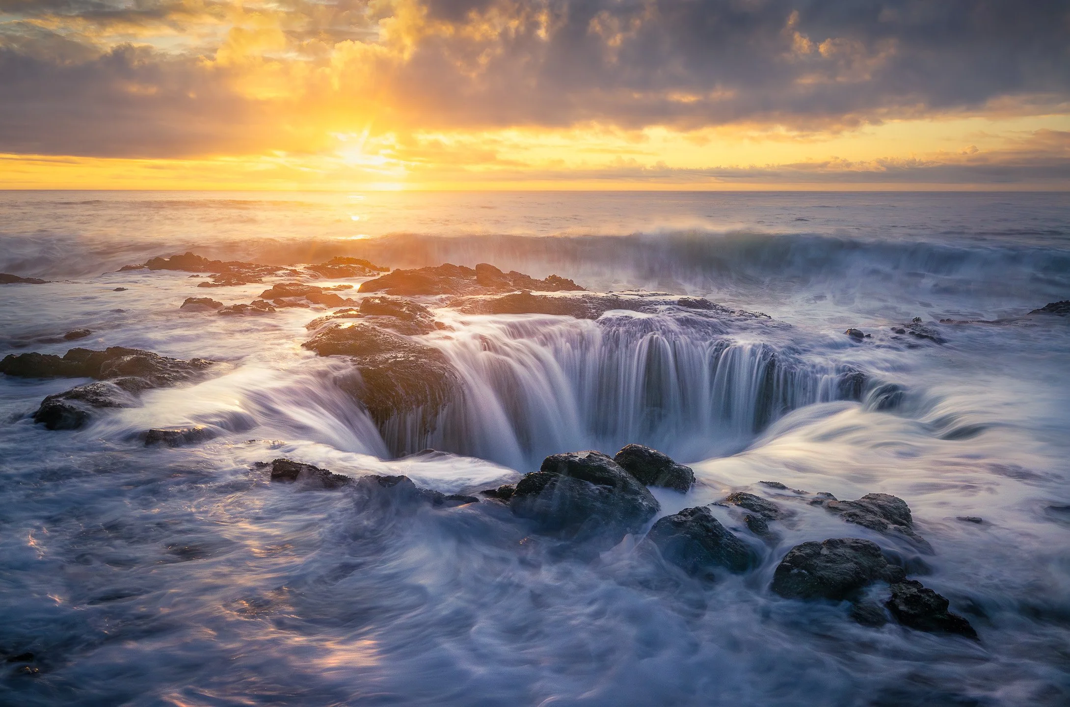 Thor's Well at Cape Perpetua during golden hour, waves crashing into the collapsed sea cave as warm sunset light colours the rocky Oregon coastline, USA.