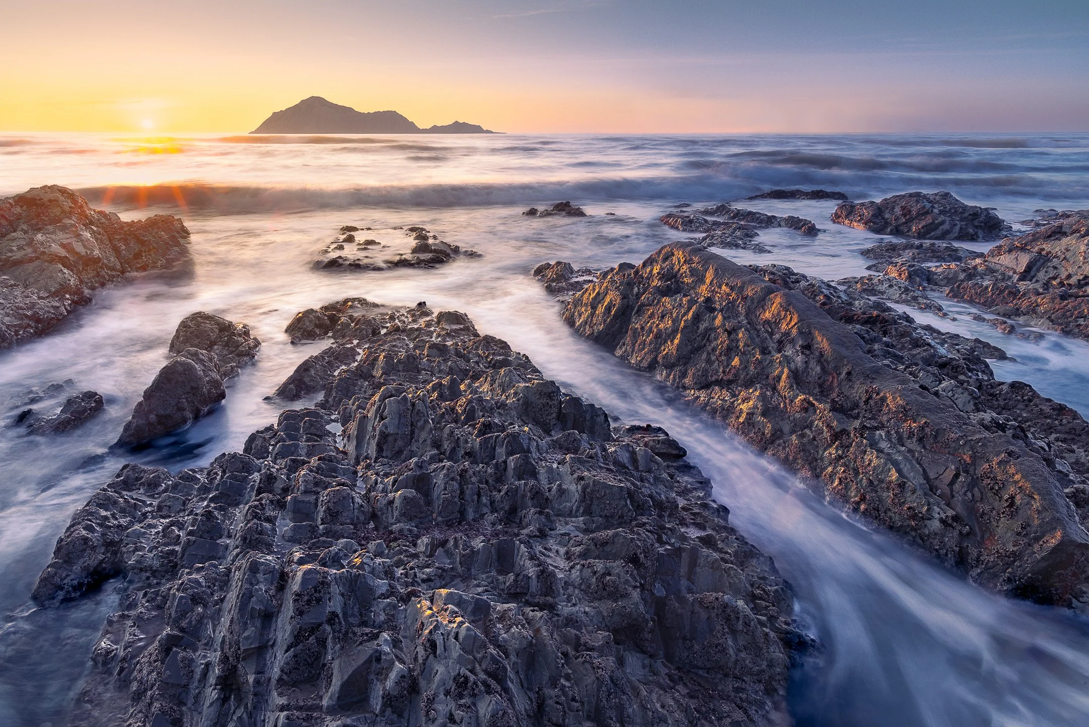 Rocky tidal ledges sculpted by the ocean at Waimārama Beach catch the light of a beautiful sunrise, with the silhouette of Bare Island visible on the horizon, Hawke's Bay, New Zealand.