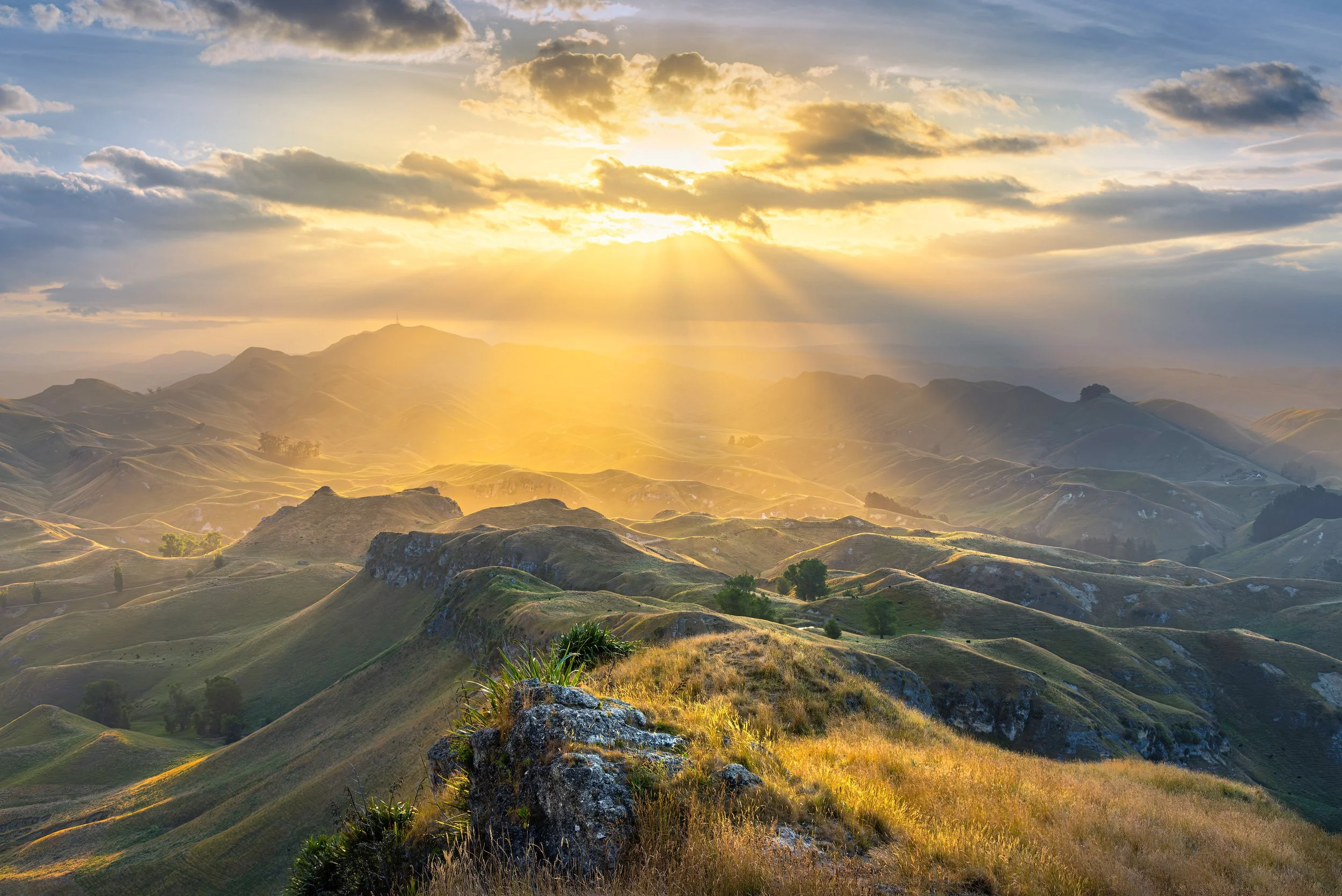 Sunbeams breaking through clouds over layered green hill country, viewed from a rocky ridge, Te Mata Park, Hawke's Bay, New Zealand.