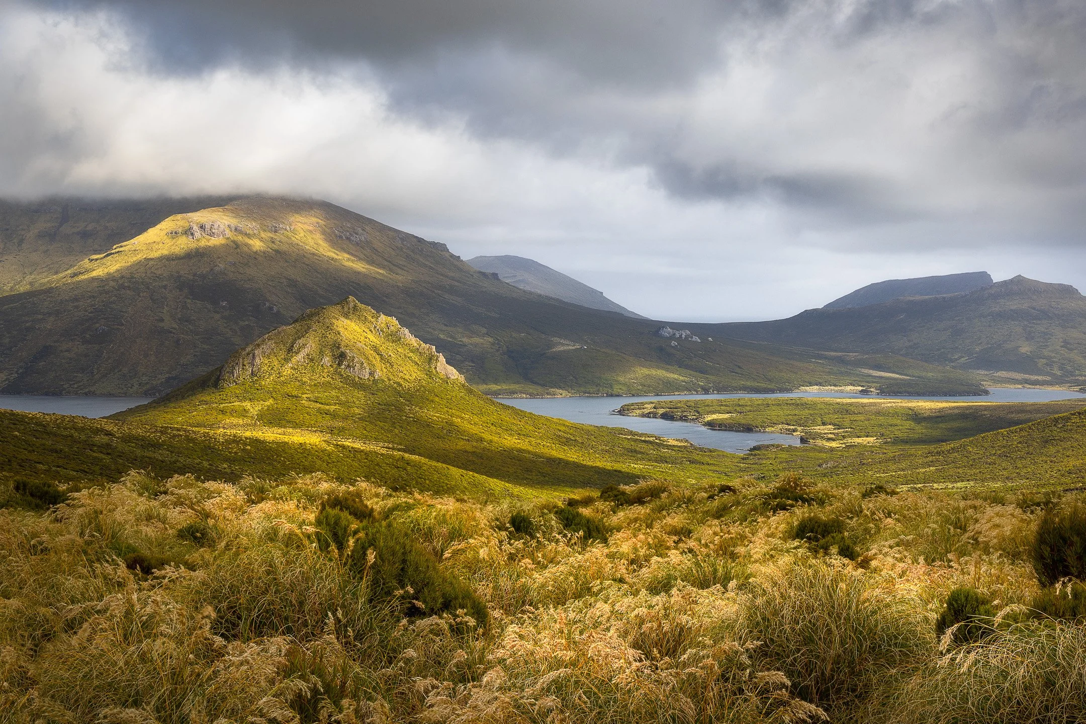 A dramatic sky above Campbell Island with broken cloud formations in grey, white, and pale blue, with tussock-covered hills and rocky terrain visible at the bottom of the frame.