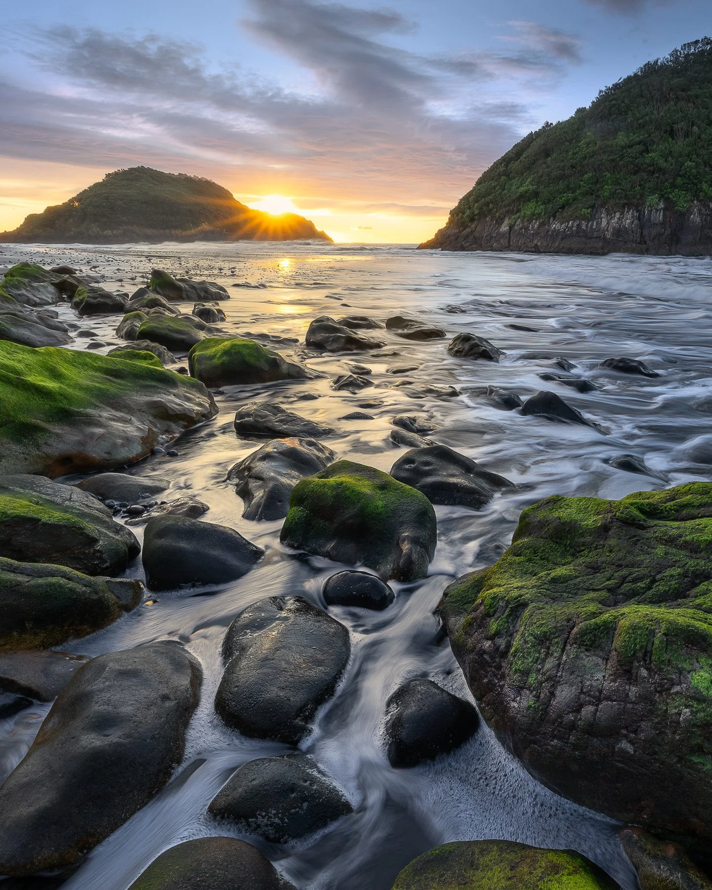 A sunstar forms over the rocky shoreline at Back Beach, New Plymouth, with two Sugarloaf Islands visible in the background, Taranaki, New Zealand.