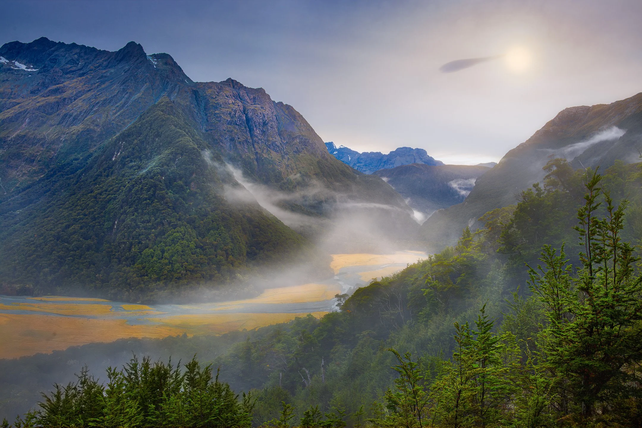 A thin veil of morning mist lingers over the Routeburn River flowing through a golden grass valley in Mount Aspiring National Park, Otago, New Zealand