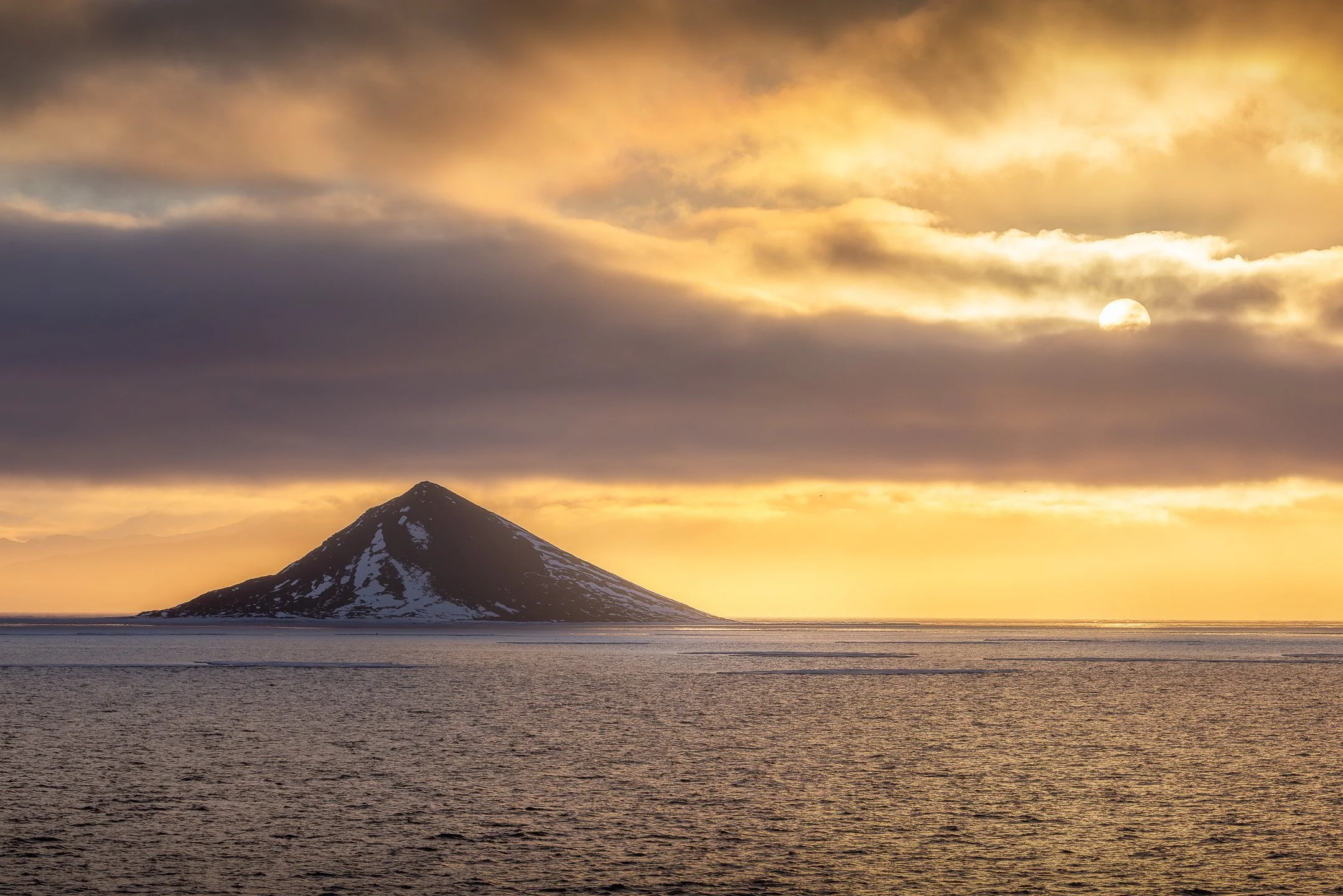 A perfectly conical volcanic peak rises from the sea, centred beneath a dramatic golden cloud system with the sun radiating behind it