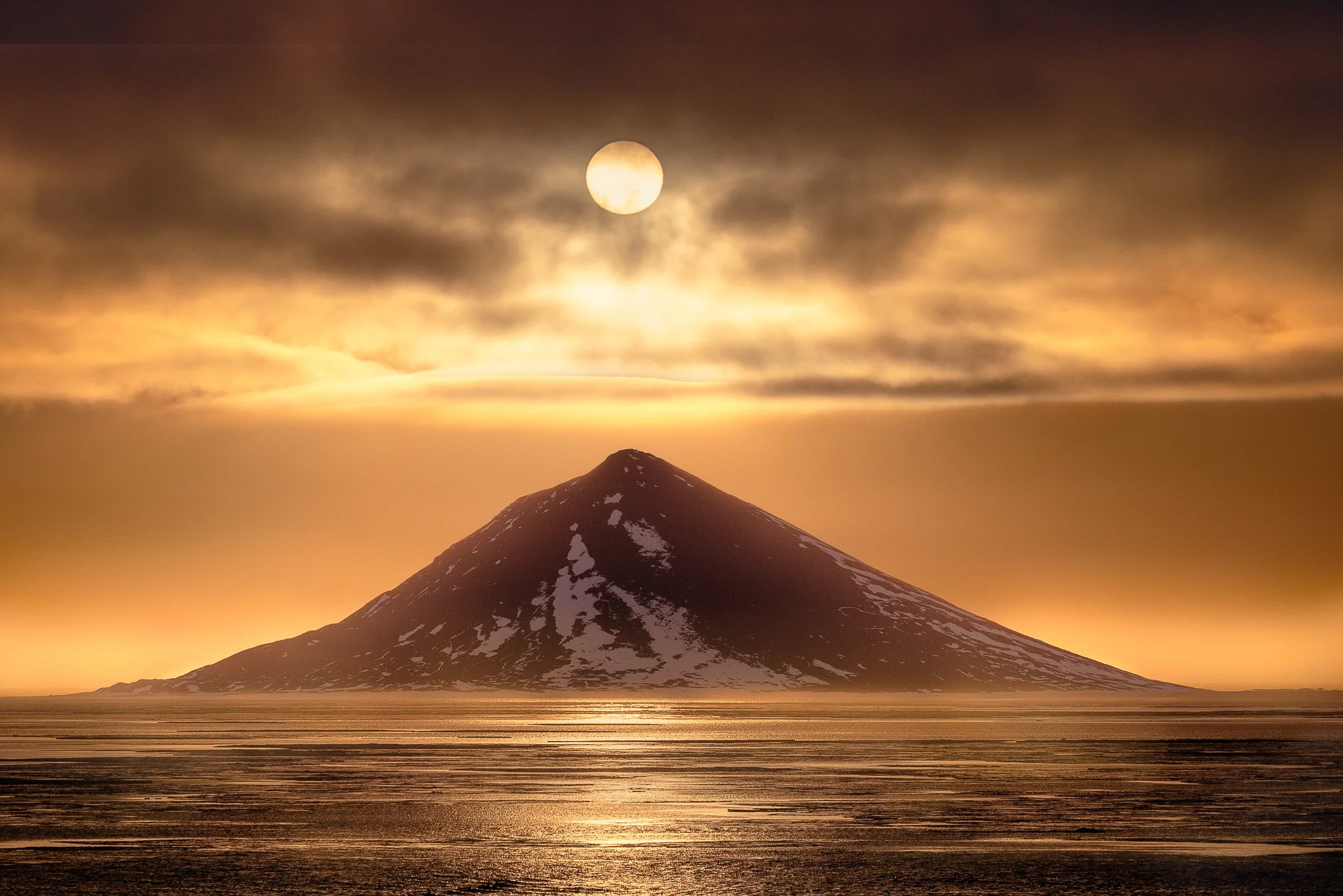 A diffused sun settles perfectly above a pyramid-shaped Antarctic island, casting a warm golden glow over the icy sea.