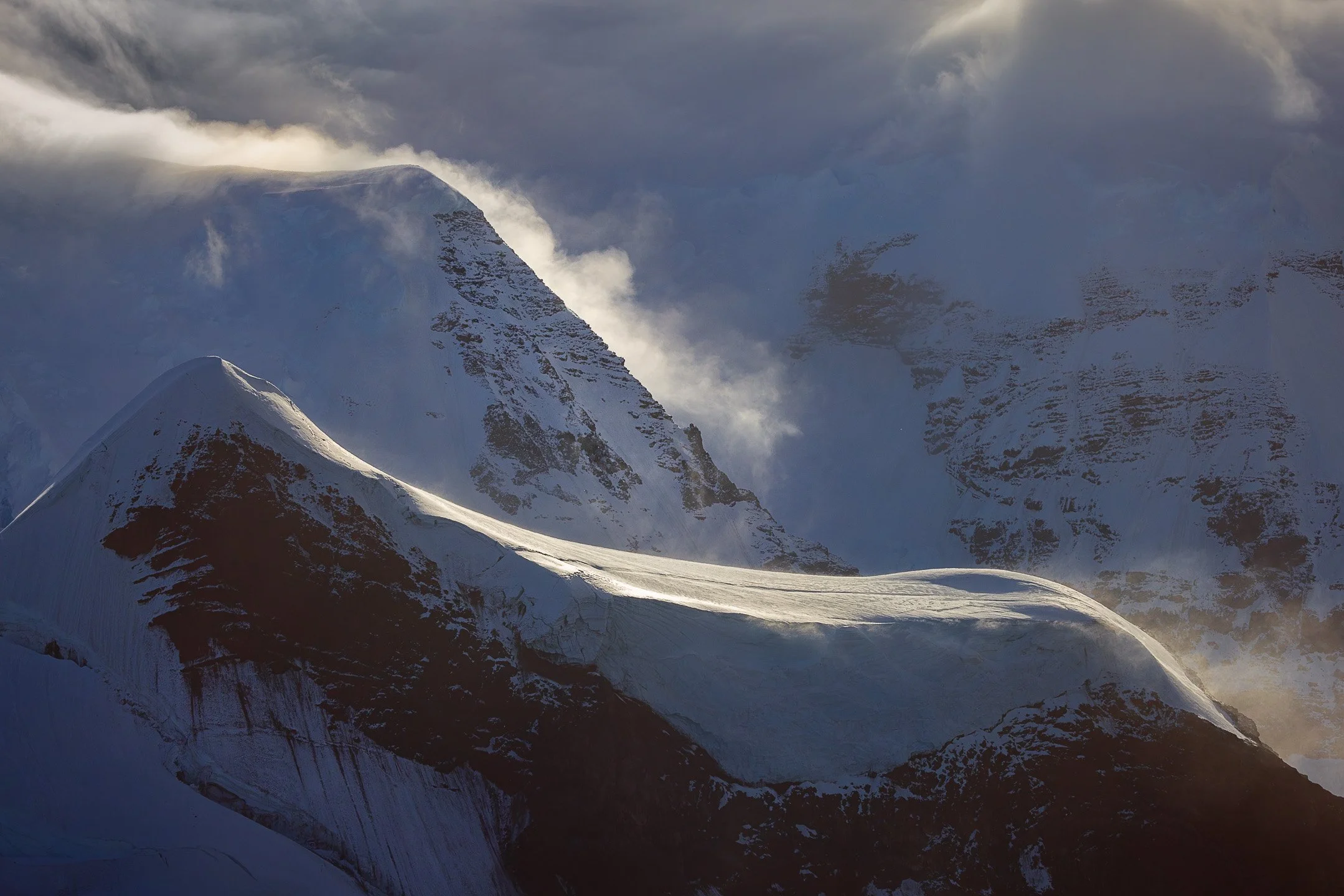 Cloud burns bright along a volcanic peak's ridgeline at the Balleny Islands, Antarctica, tracing its edge against the sky while a curve of glacial ice catches the same fierce directional light below.