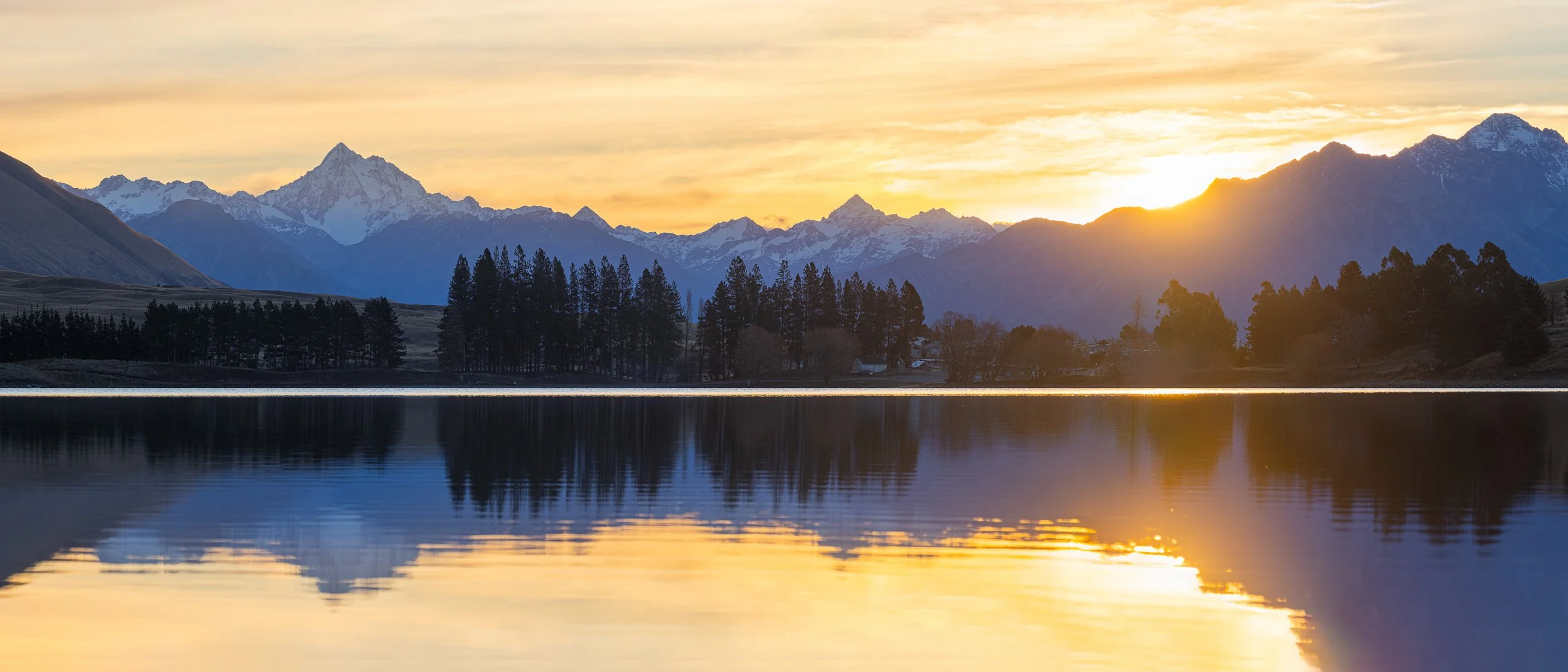 Sunset reflection of Mt D'Archiac, McClure Peak, Cloudy Peak and Mount Potts in Lake Camp, Ashburton Lakes, Canterbury, New Zealand, with pine trees and snow-capped Southern Alps mirrored in still water.