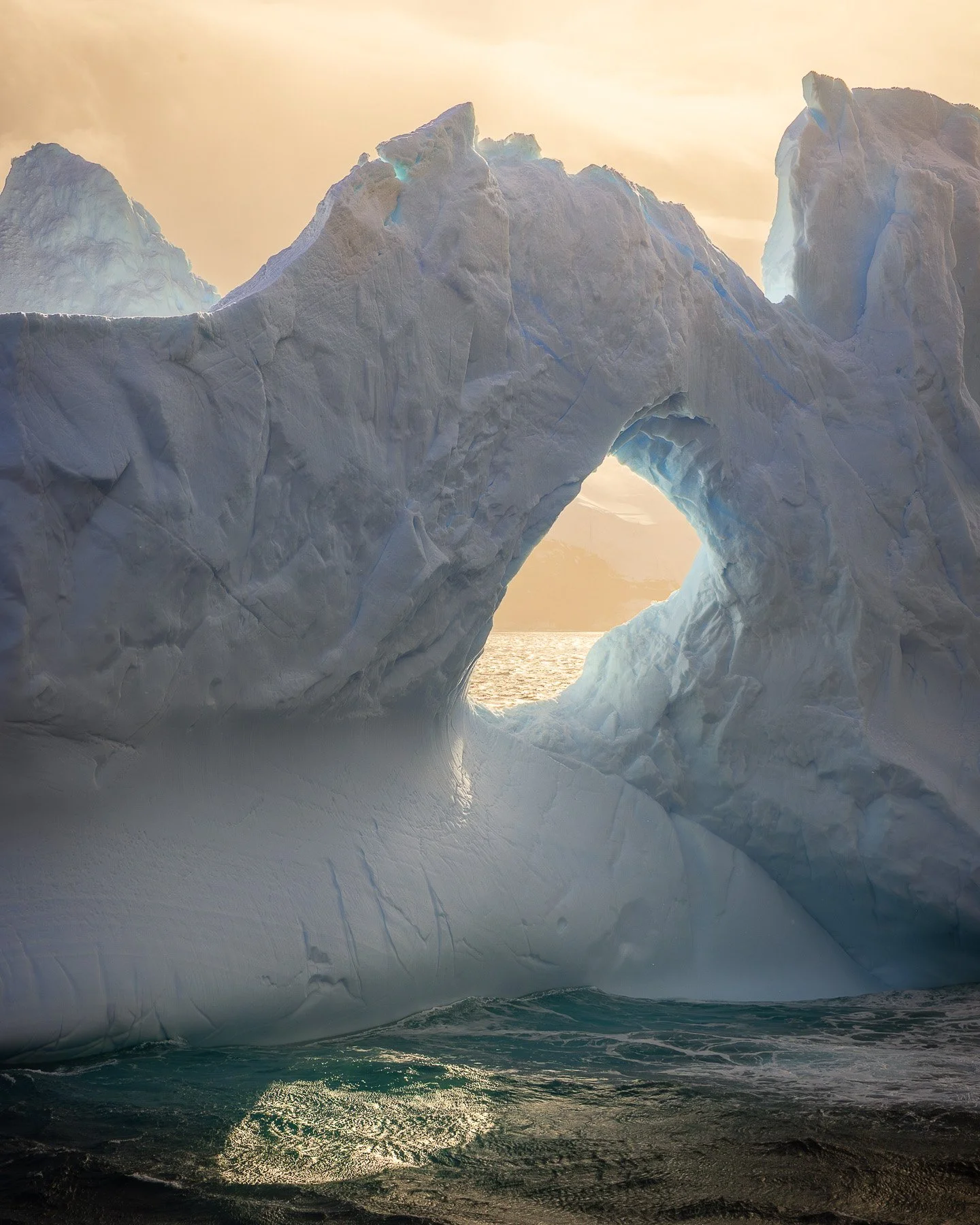 Looking up and through a monumental ice arch near the Balleny Islands, Antarctica — golden sky framed by sculpted ice, the Southern Ocean visible at the edges of the opening.