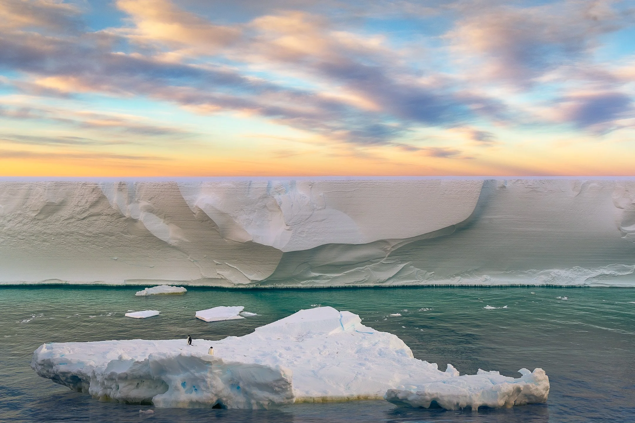 Two Adélie penguins stand on a small irregular ice floe drifting past the vast white face of the Ross Ice Shelf, Ross Sea. The water between the floe and the shelf is teal-green, and the sky above shows bands of gold, blue, and lavender at sunset.