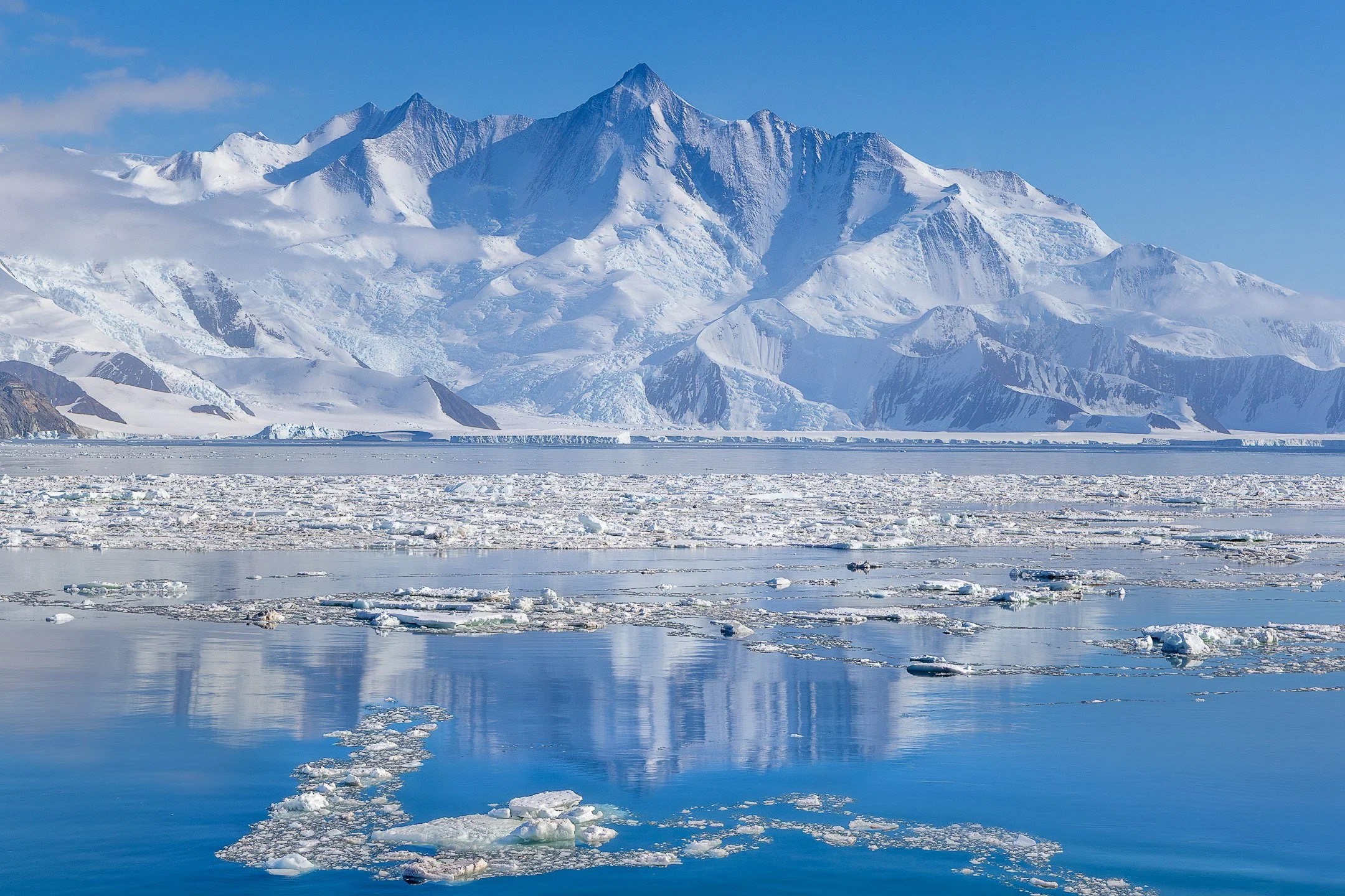Brindled ice and sea ice fragments float in a calm Antarctic bay, reflecting a massive, jagged mountain range under a clear sky.