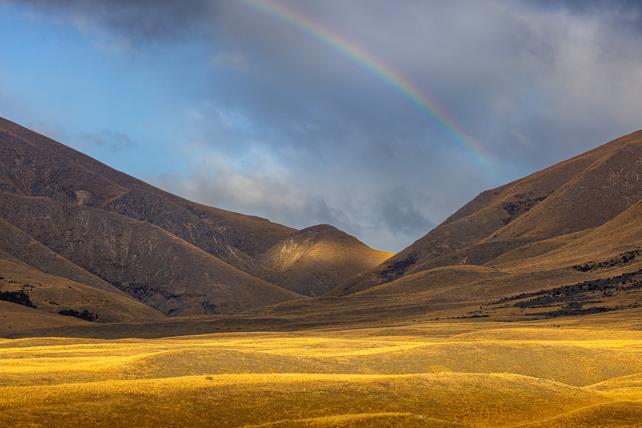 Rainbow fragment descending into storm clouds above a mountain valley, with a patch of golden sunlight on the central saddle and warm tussock grassland in the foreground, Lake Camp, Canterbury.