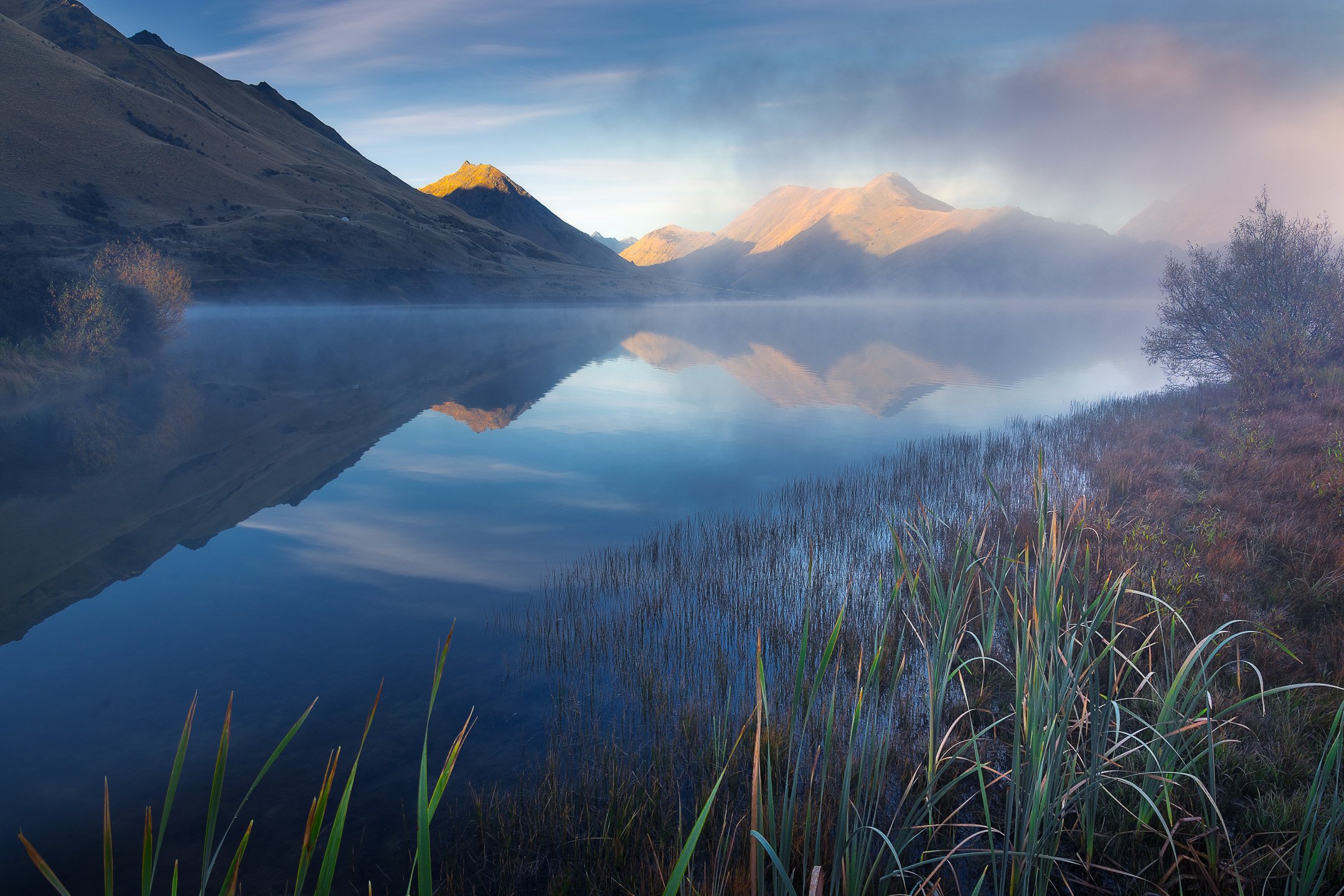Morning fog lifts slowly from the mirror-like surface of Moke Lake near Queenstown, the surrounding mountain peaks gradually revealing themselves through retreating mist, Otago, New Zealand.