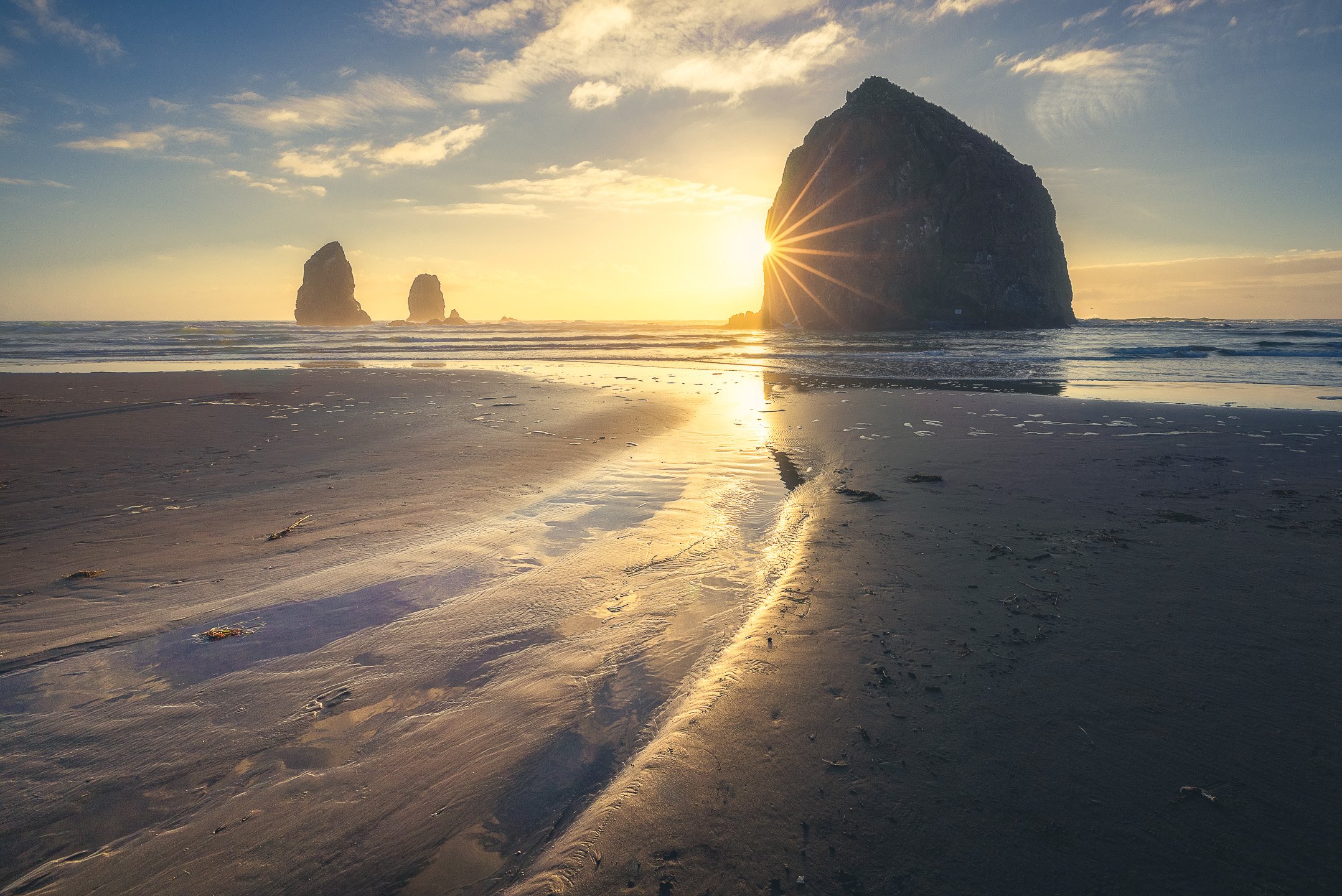 Haystack Rock at Cannon Beach during golden hour, a sunstar forming beside the towering formation as a tidal stream curves across the sand toward the rock, Oregon, USA.