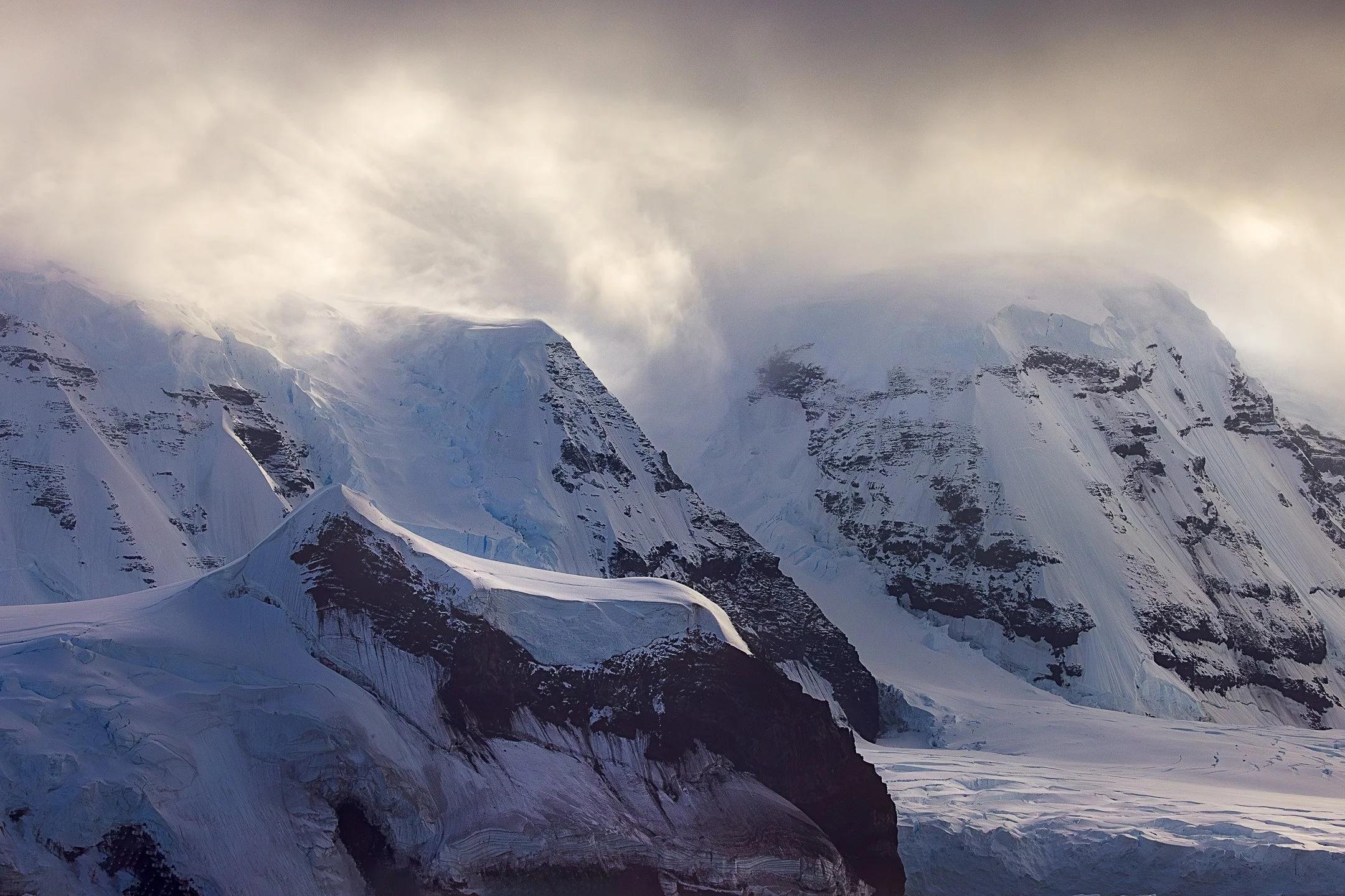 Volcanic peaks of the Balleny Islands sit beneath a pressing cloud ceiling, their dark rock faces and glacial flanks caught in the charged atmosphere between storm and sky.