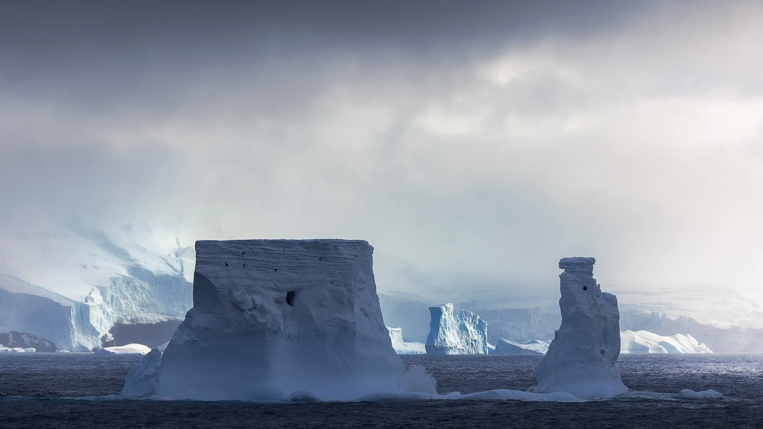 Two carved ice forms frame the entrance to the Balleny Islands in the Southern Ocean, with icebergs, cliffs and glaciers stretching away into the mist beyond them.