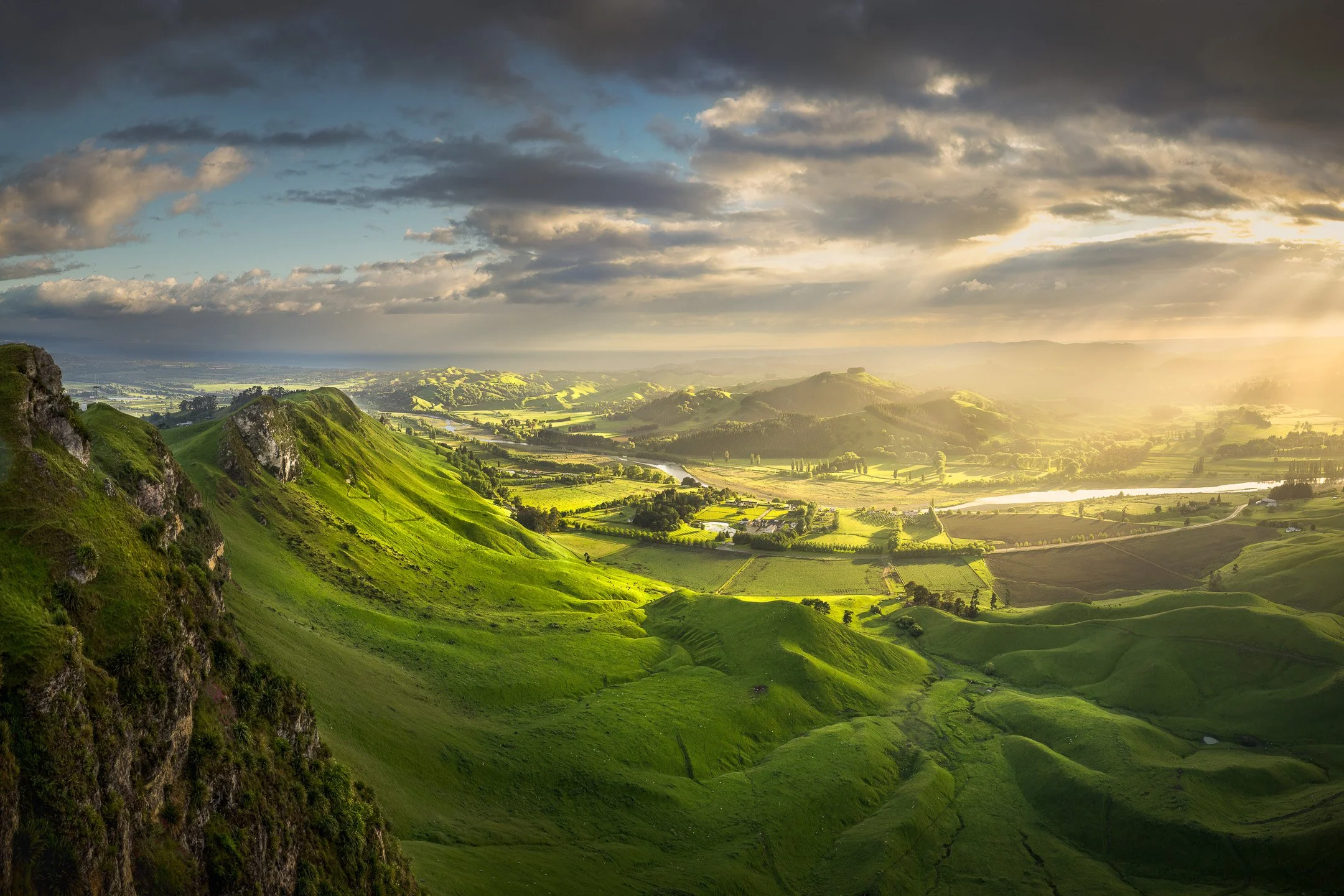 Aerial view from Te Mata Peak at sunrise, with sun rays breaking through cloud over a green river valley and rolling farmland in Hawke's Bay, New Zealand.
