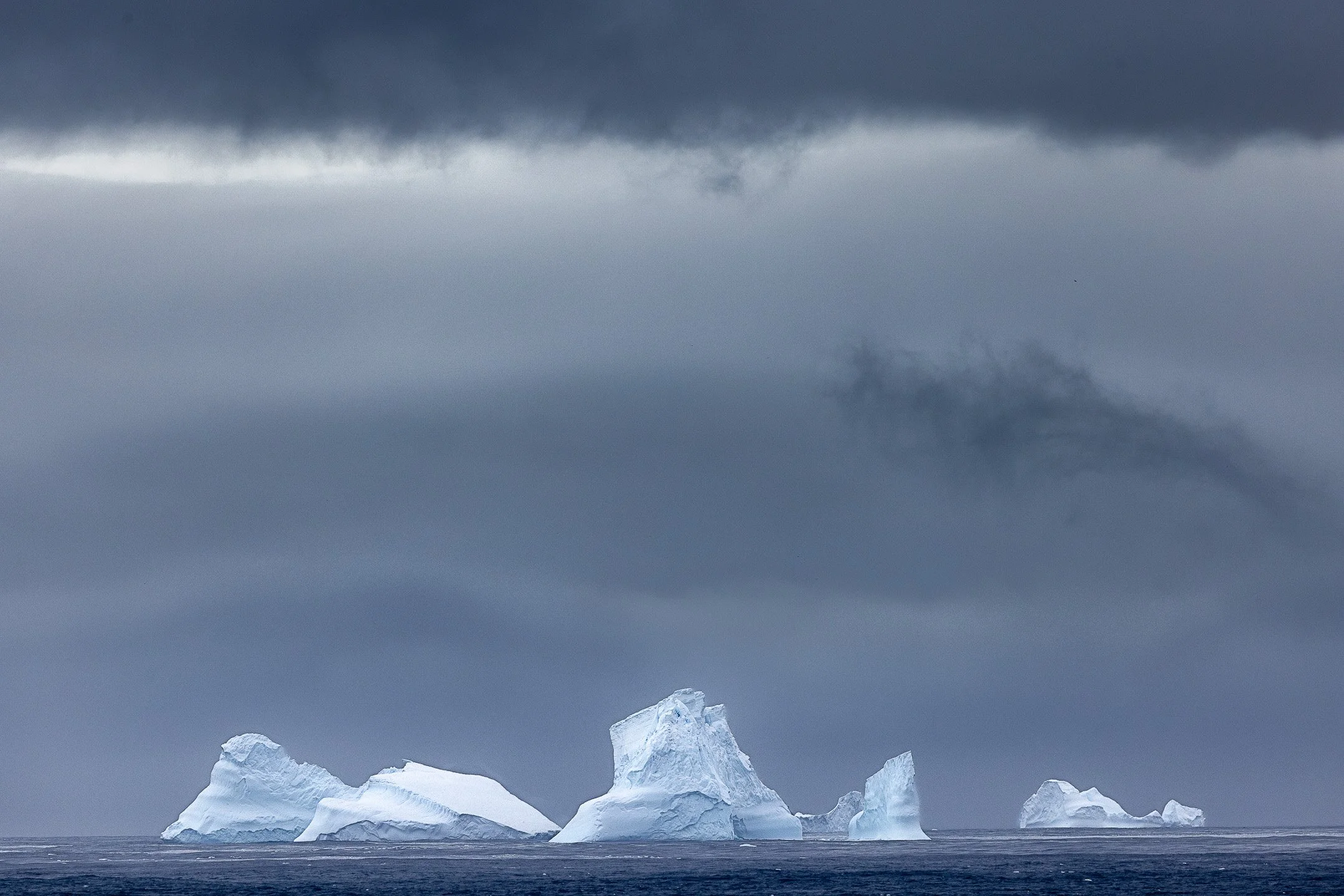 A scattered group of icebergs sits low on the horizon beneath a heavy layered sky in the Southern Ocean near the Balleny Islands, Antarctica.