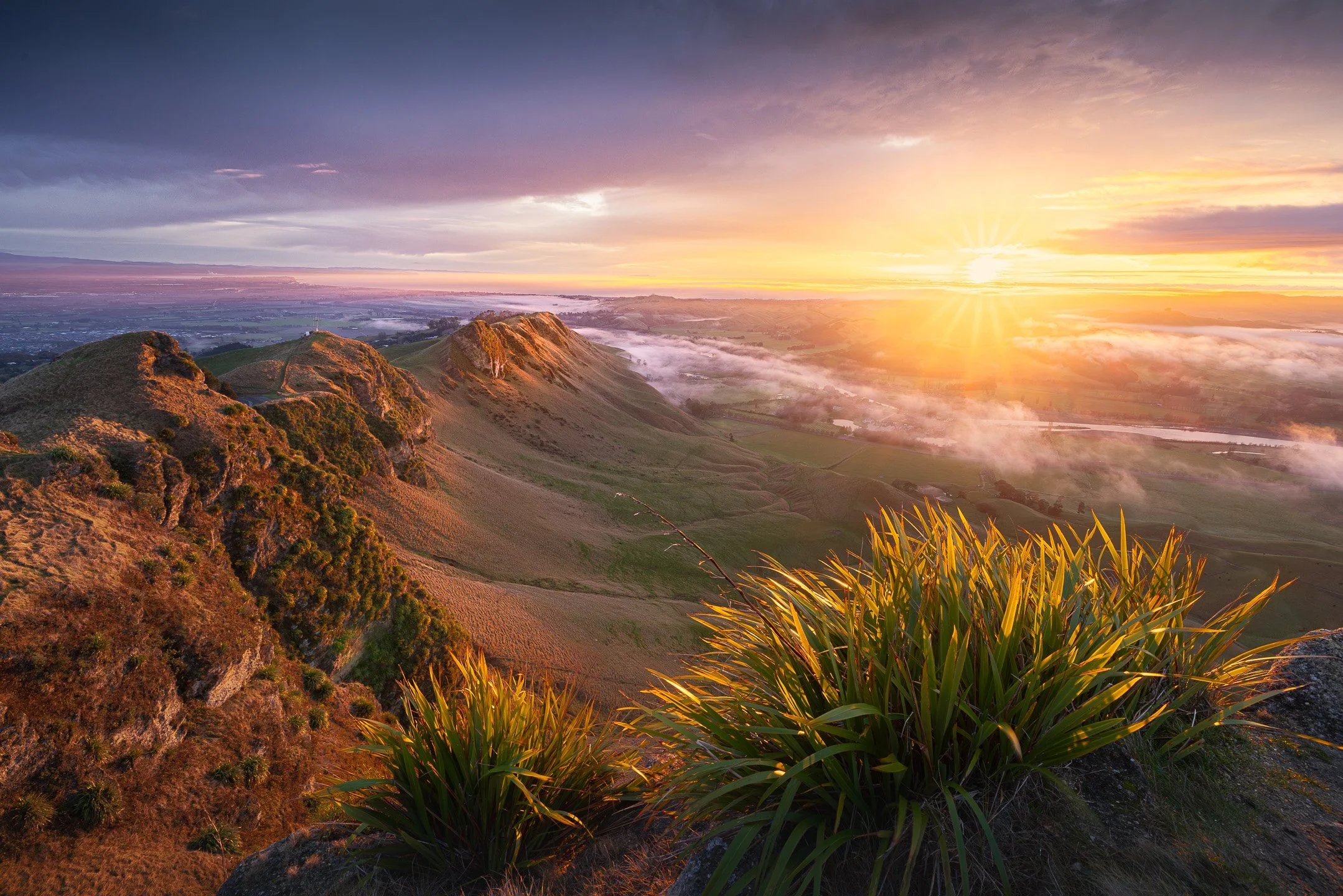 Warm golden sunrise light bathes Te Mata Peak as scattered fog drifts above the ocean and across the Hawke's Bay landscape on a winter morning, New Zealand