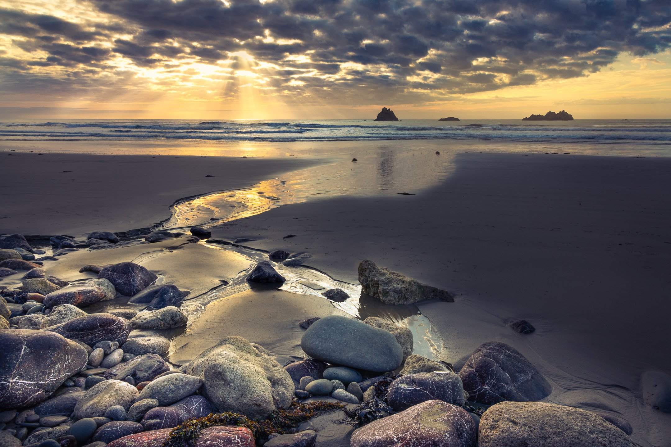 A tidal stream leads the eye toward the Hinemahanga Rocks at Kairakau Beach as sunbeams break through rolling clouds above, Hawke's Bay, New Zealand.