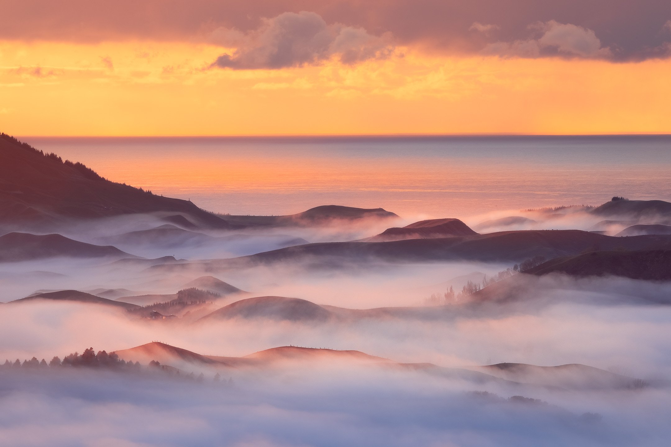 Golden sunrise over mist-covered hills in Hawke's Bay, New Zealand, with low cloud filling the valleys and the Pacific Ocean visible on the horizon.