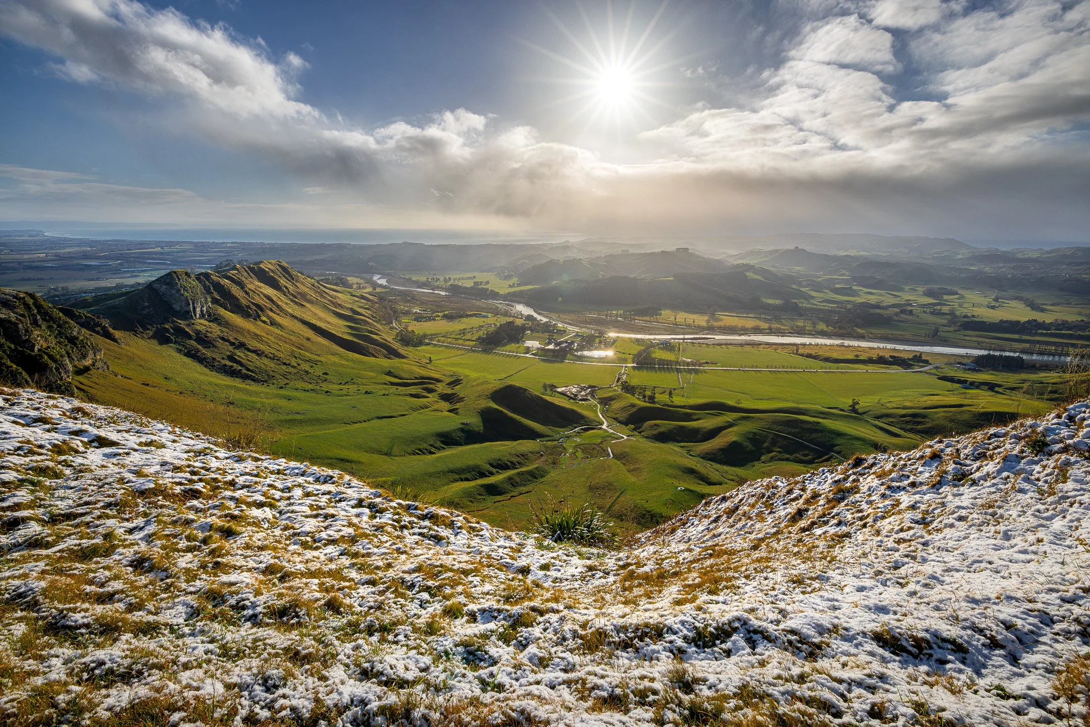 Sunburst breaking through clouds over a snow-dusted Te Mata Peak, with a river winding through the green valley below.