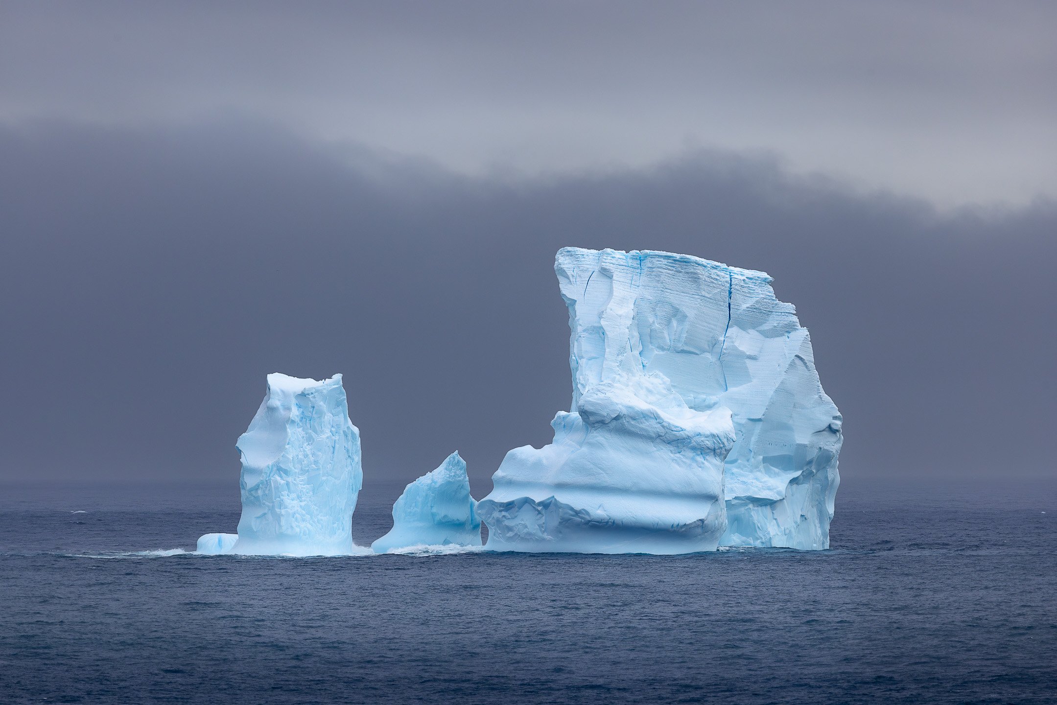Two icebergs in close proximity drift through the Southern Ocean near the Balleny Islands — one massive and blocky, one smaller and more delicate, both luminous against the steel-grey sea.
