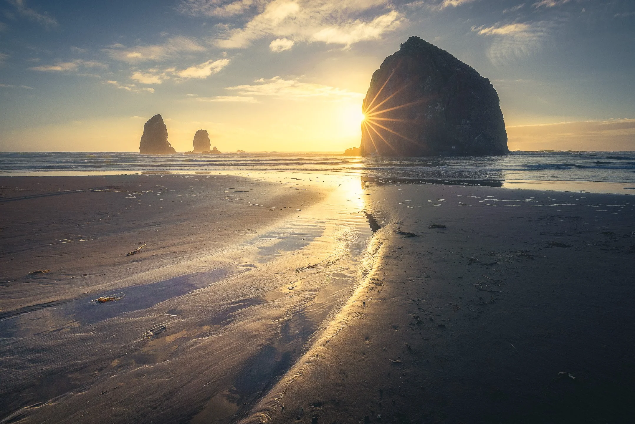 Haystack Rock at Cannon Beach during golden hour, a sunstar forming beside the towering formation as a tidal stream curves across the sand toward the rock, Oregon, USA.