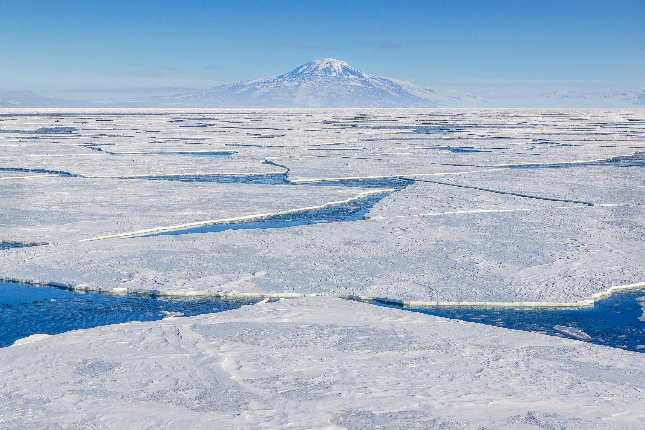 Huge cracked ice floes of the Antarctic sea ice lead toward a lone, snow-covered volcanic peak under a clear blue sky.